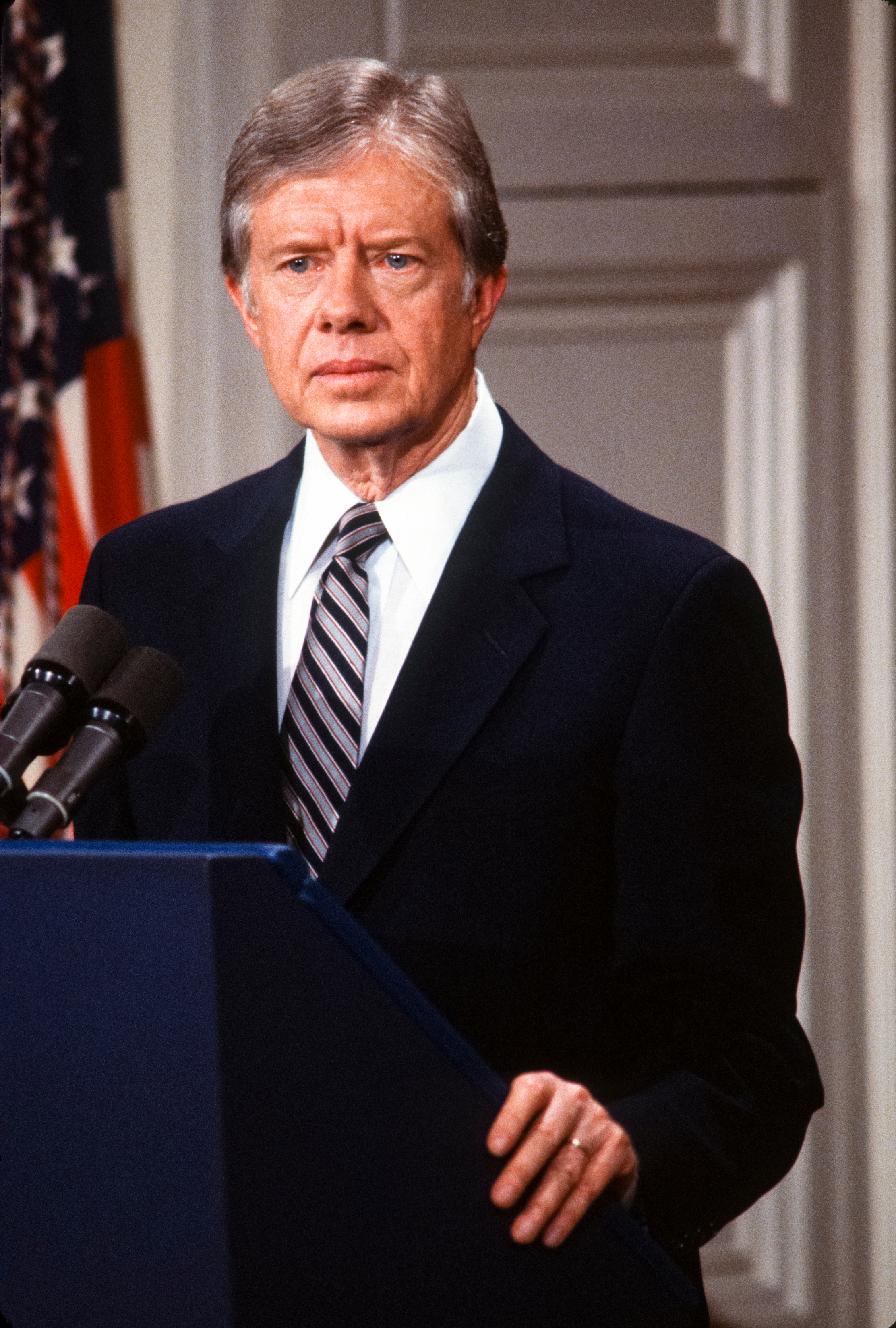 Jimmy Carter standing at a podium giving a speech, wearing a suit and tie, with the American flag in the background