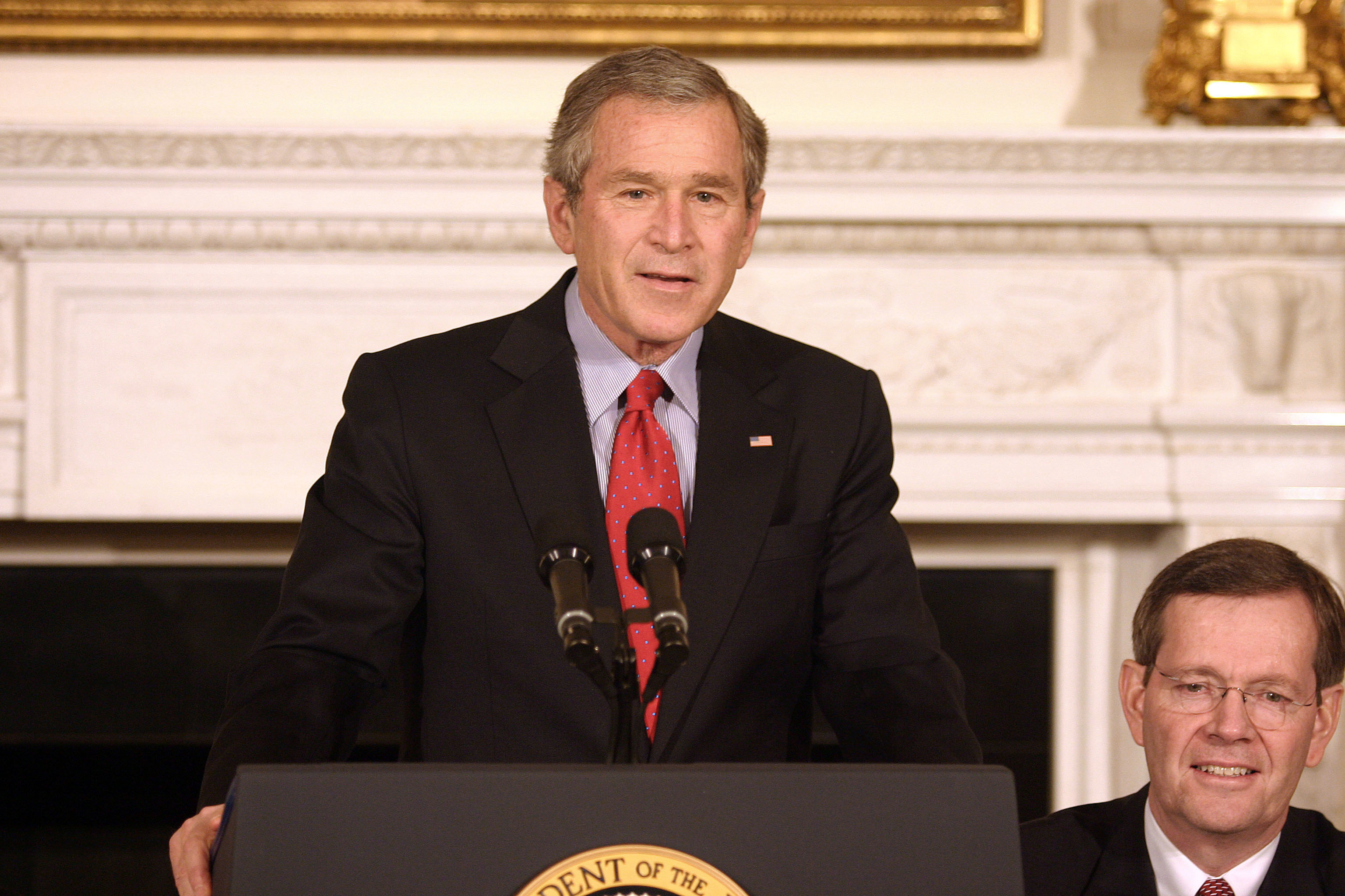 George W. Bush speaks at a podium with the Presidential Seal. A man in glasses and a suit is seated to his right