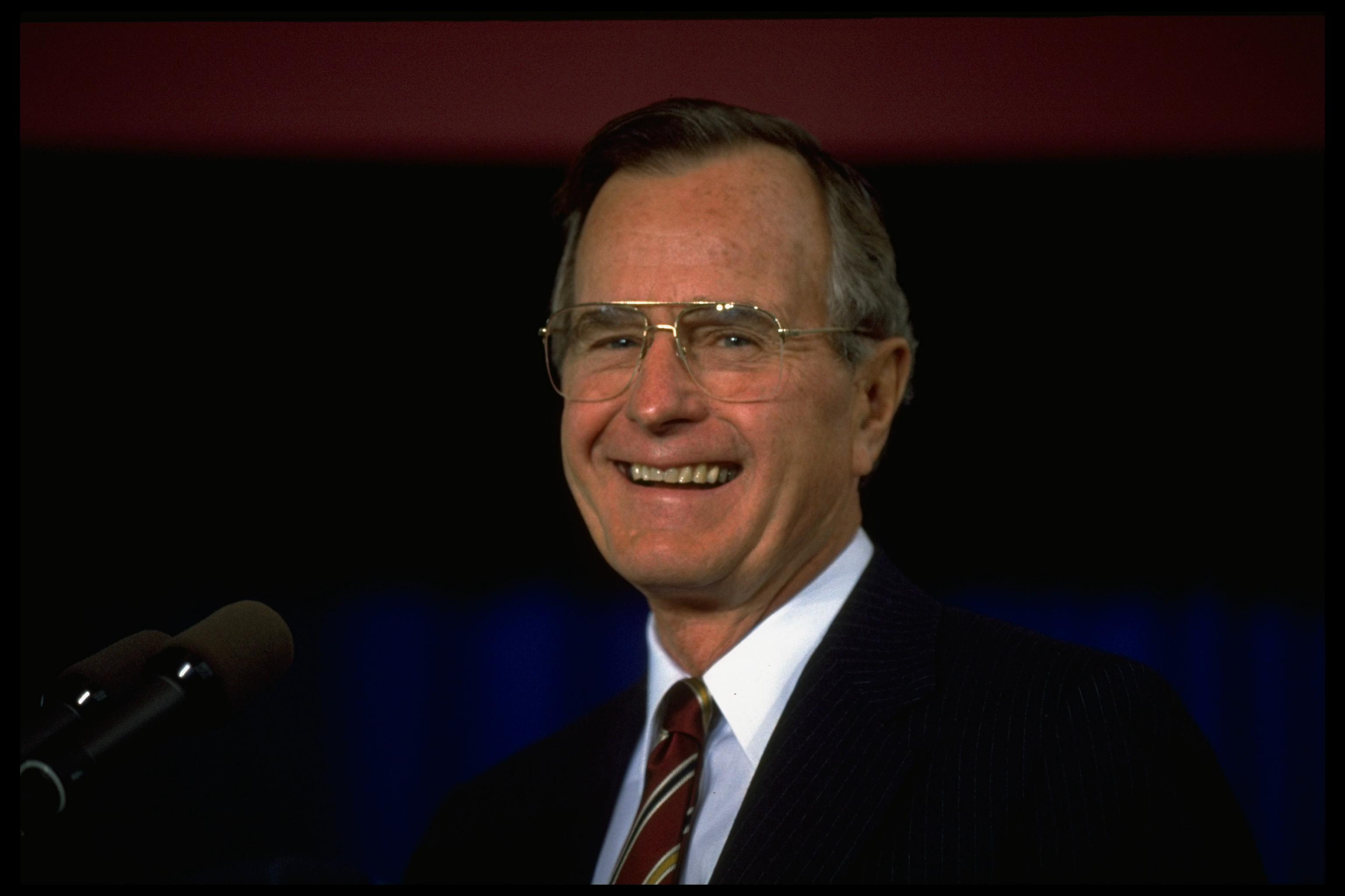 George H.W. Bush smiles while wearing a suit and tie, standing in front of a microphone