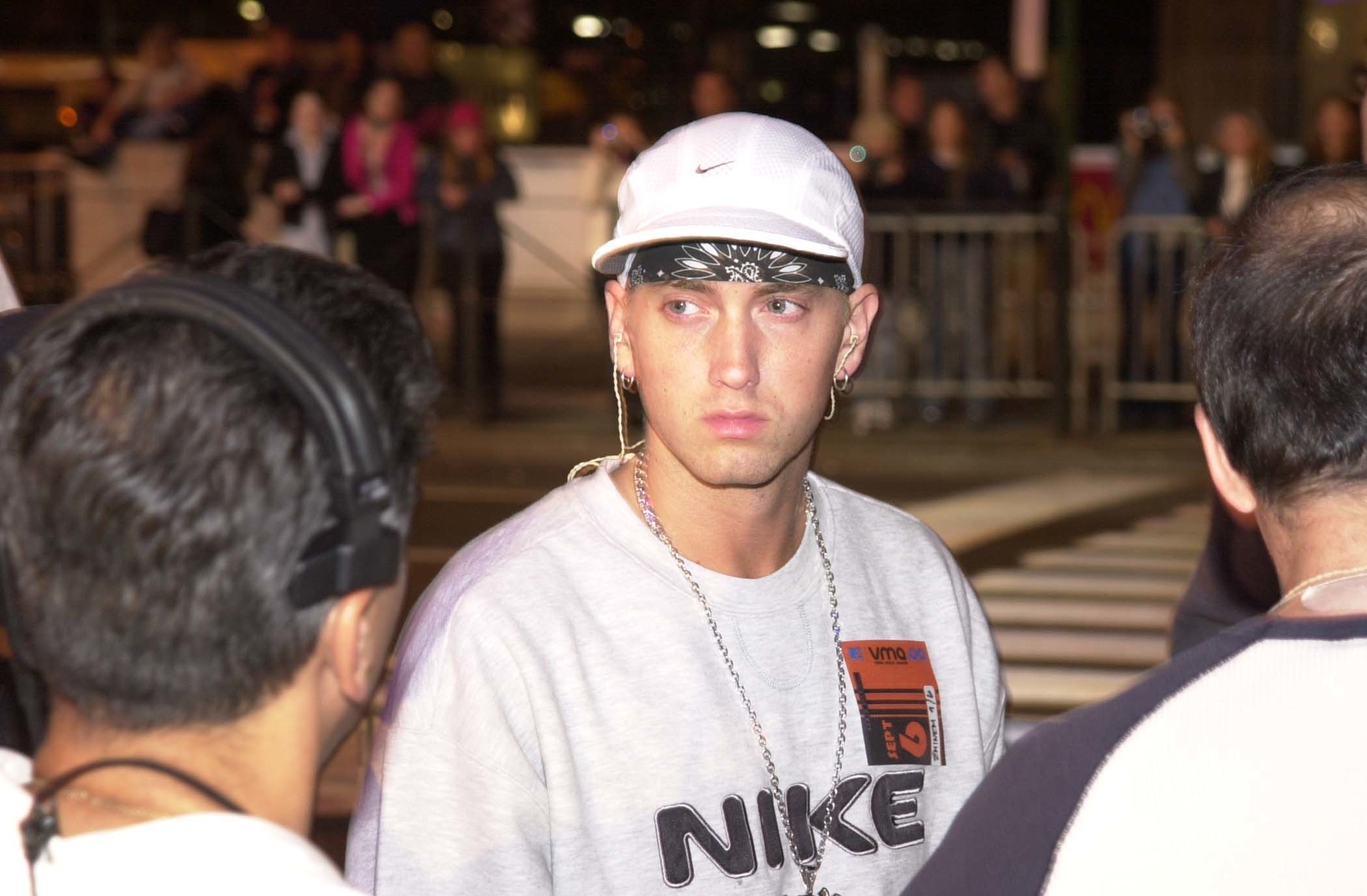 Eminem stands on a street wearing a white Nike cap, a bandana, a gray Nike sweatshirt with a small logo, and a chain necklace. People are visible in the background