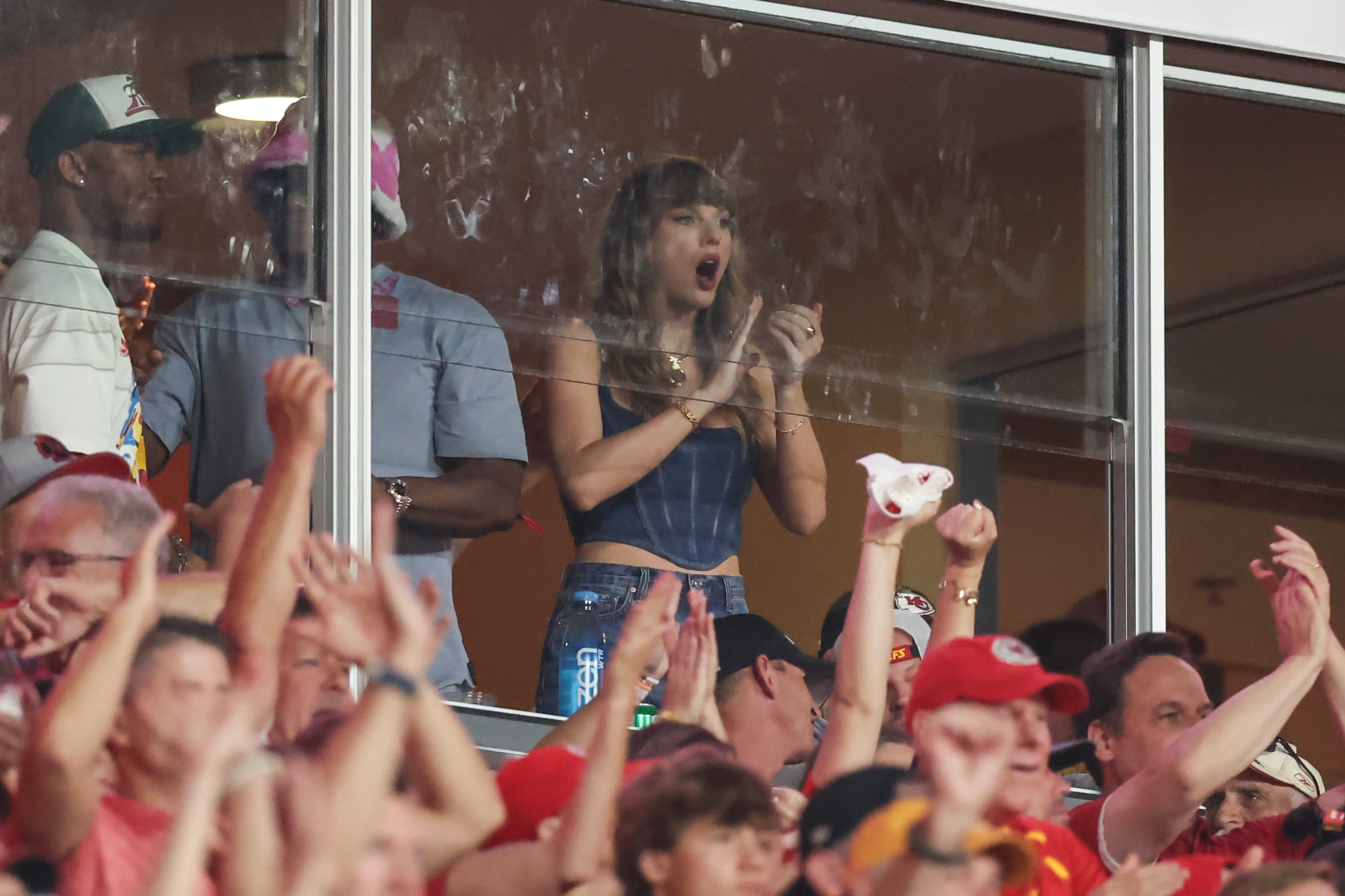 Taylor Swift cheers enthusiastically from a glass-enclosed box at a sports event, surrounded by many fans