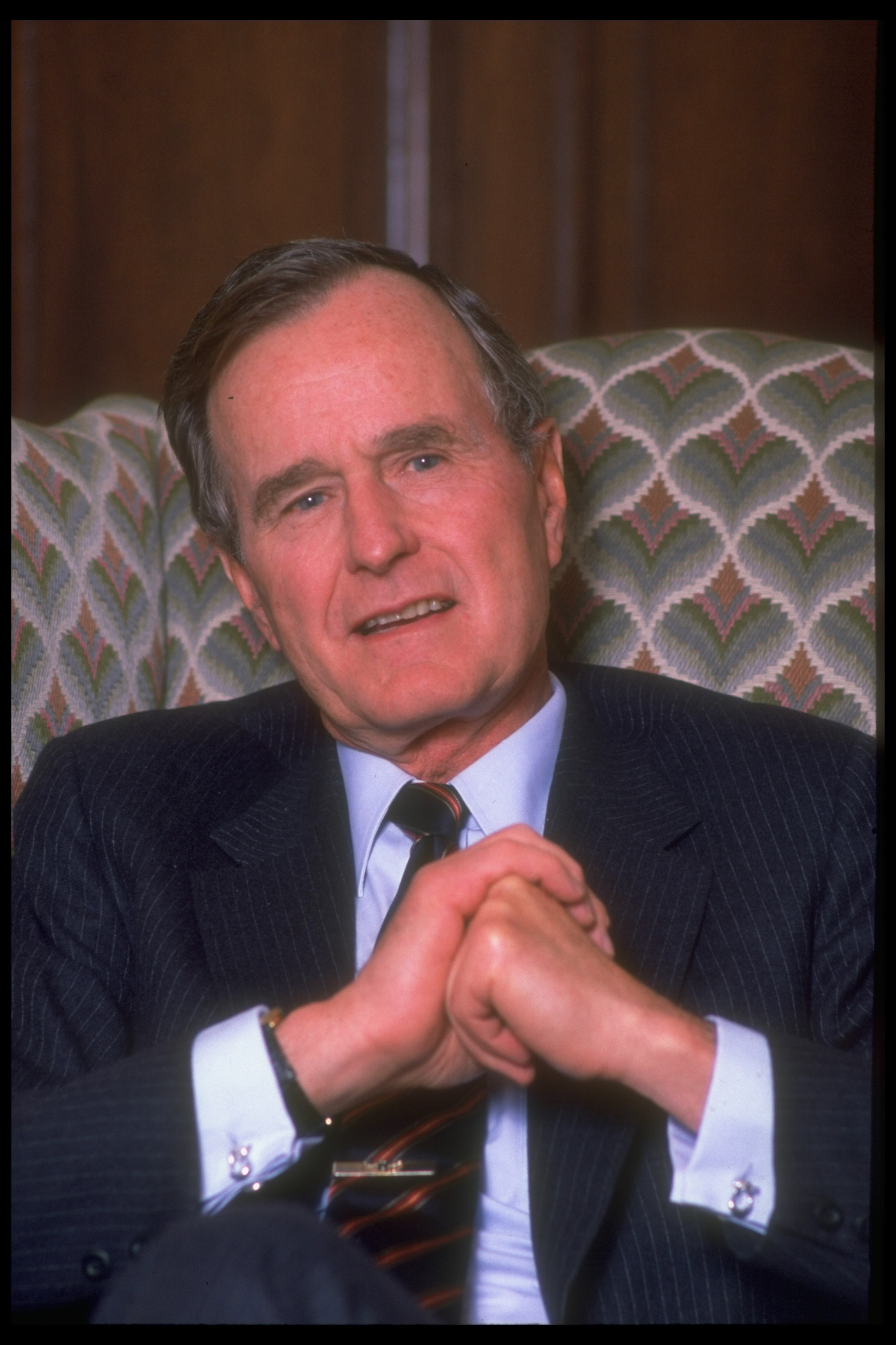 George H.W. Bush, wearing a dark suit and tie, sits in a patterned armchair, hands clasped
