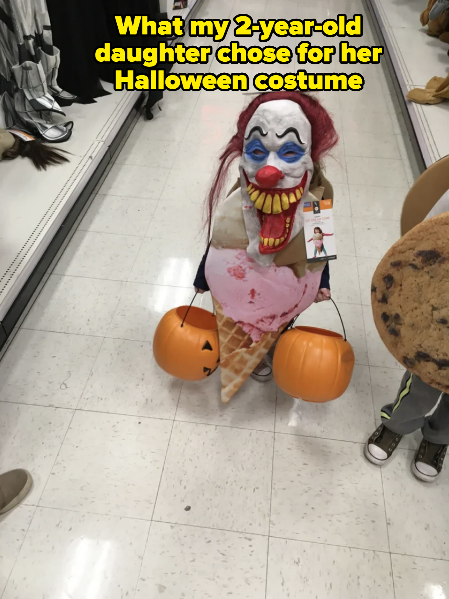 A child in a creepy clown mask with red hair and a large smile holds two orange pumpkin buckets. They wear an ice cream cone costume with a pink scoop on top