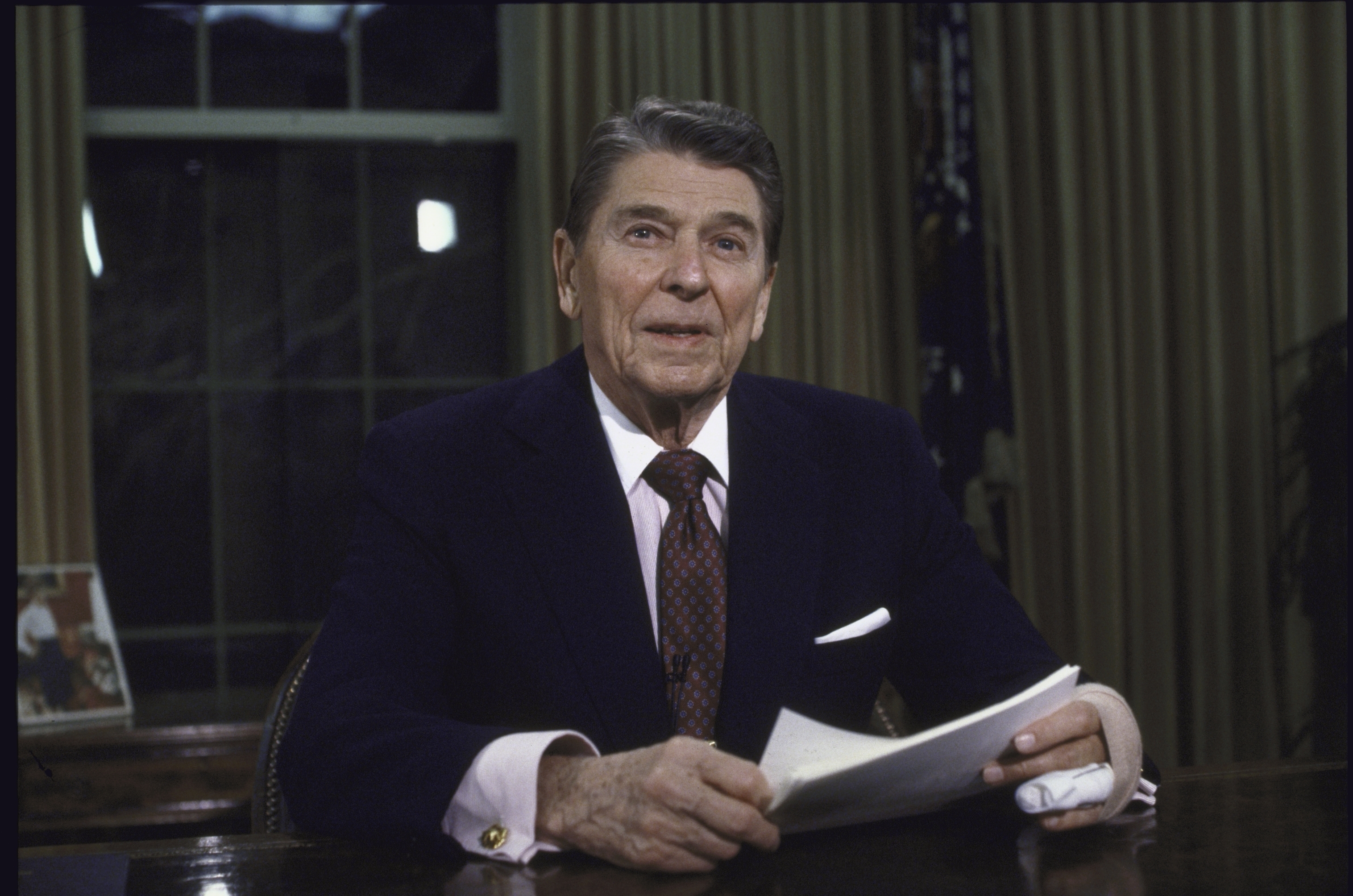 Ronald Reagan sits at a desk in an office, holding documents and smiling at the camera