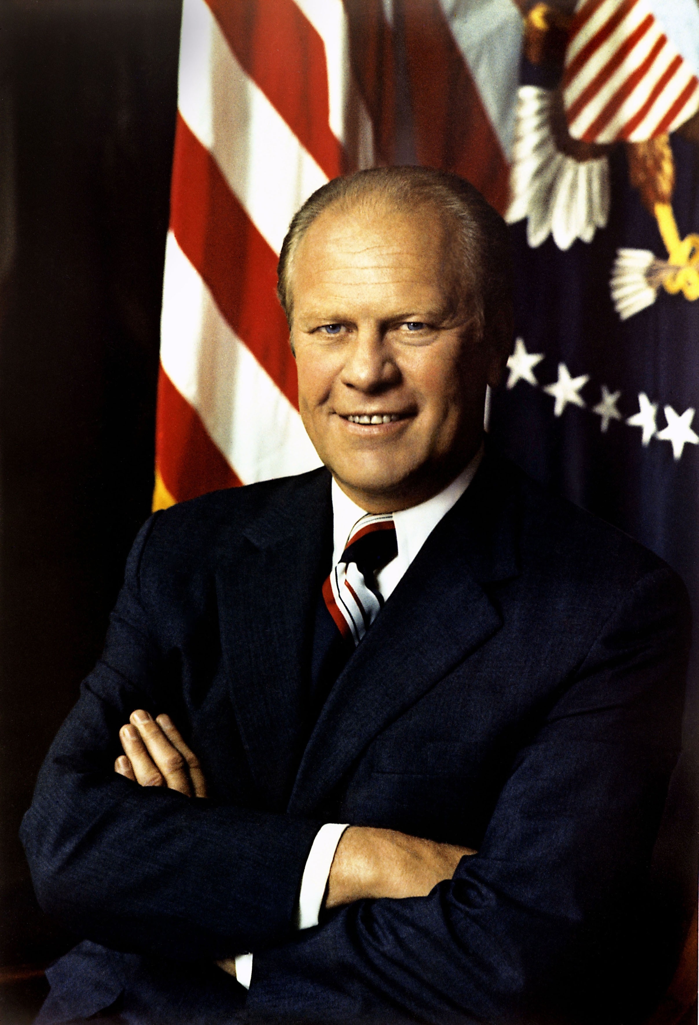 Gerald Ford, in a suit and tie, stands with arms crossed in front of U.S. flags and presidential seal