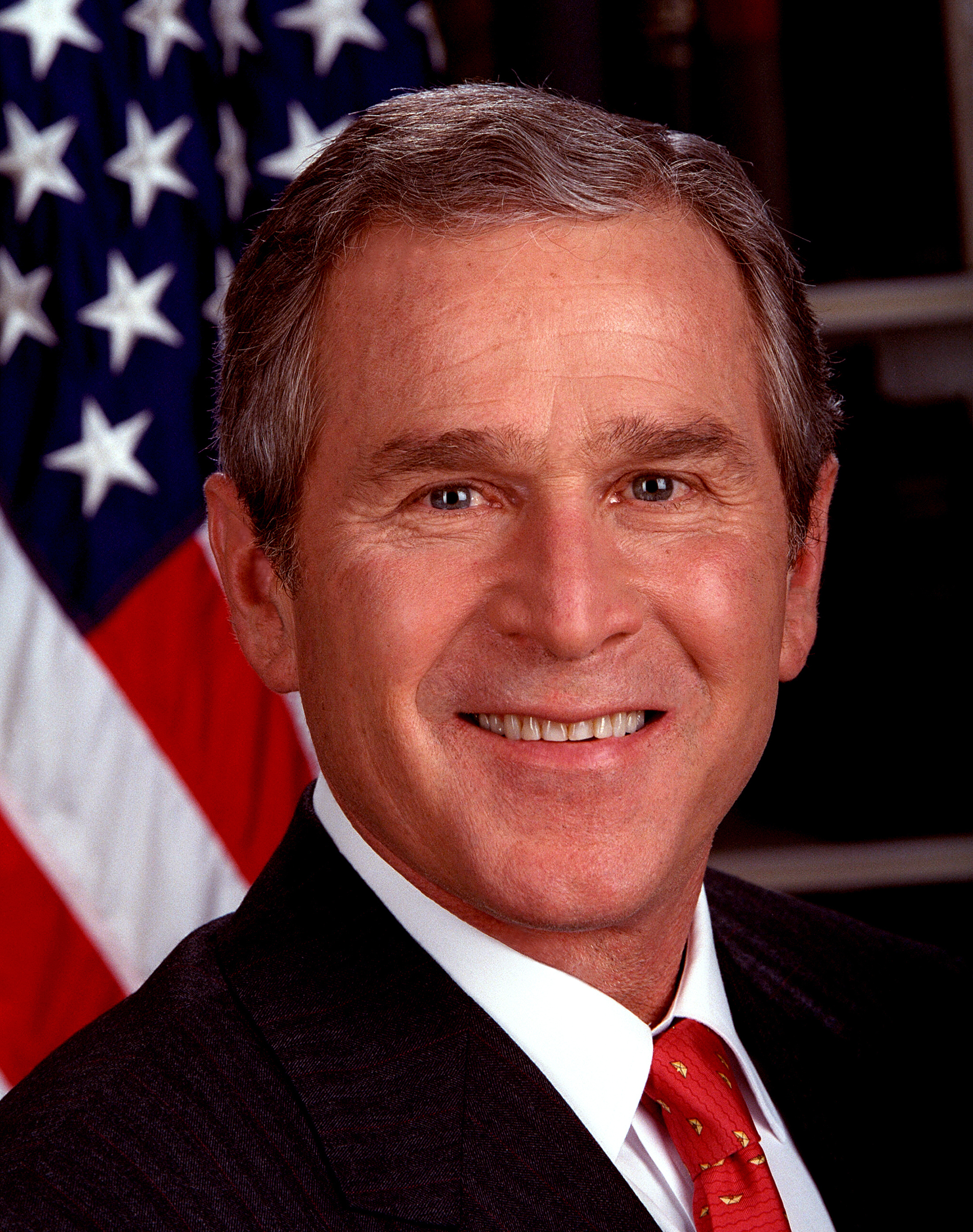 A smiling person in a suit and red tie is posing in front of an American flag