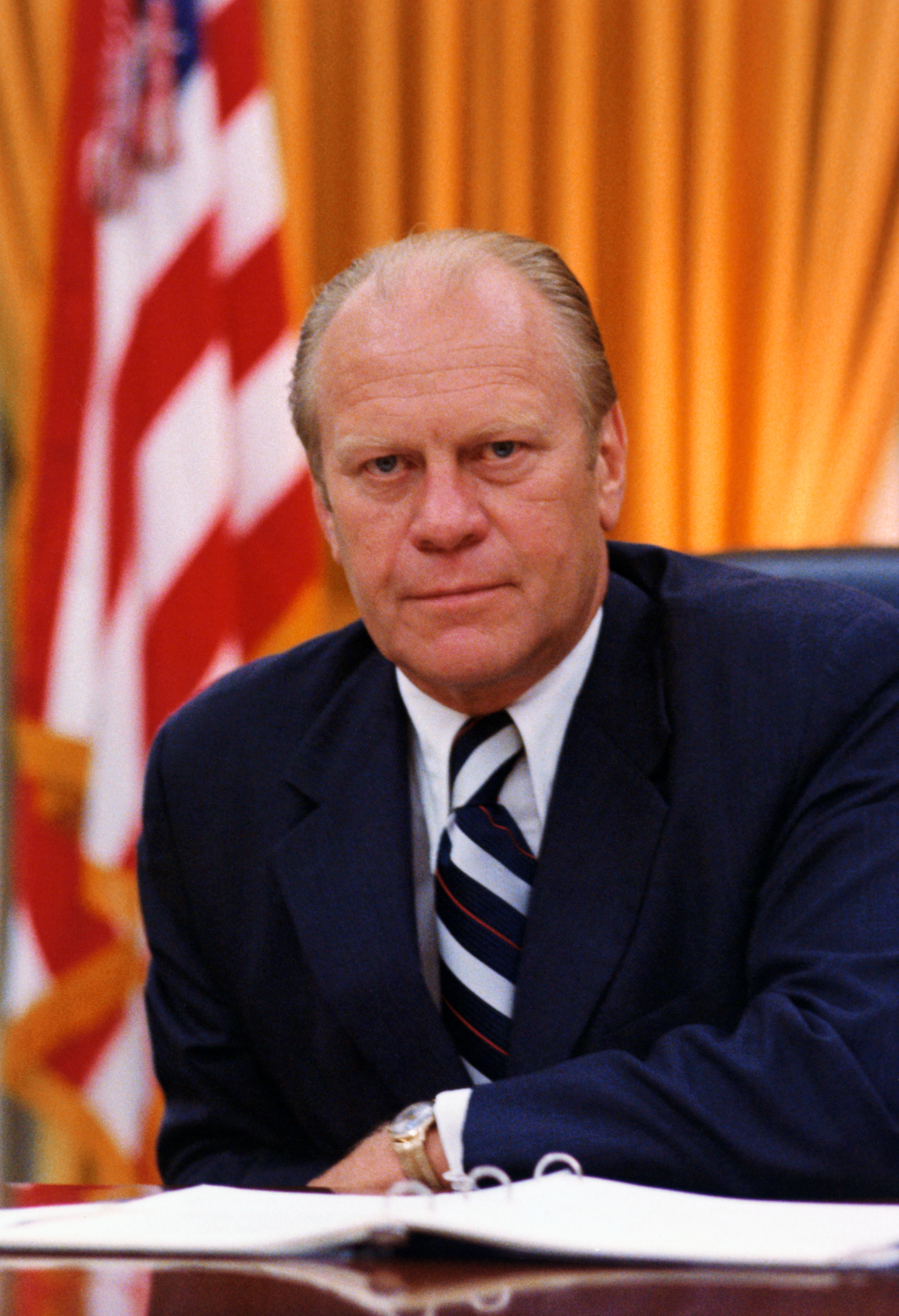 Gerald Ford sits at a desk in an office with an American flag and curtain in the background. He wears a suit and striped tie