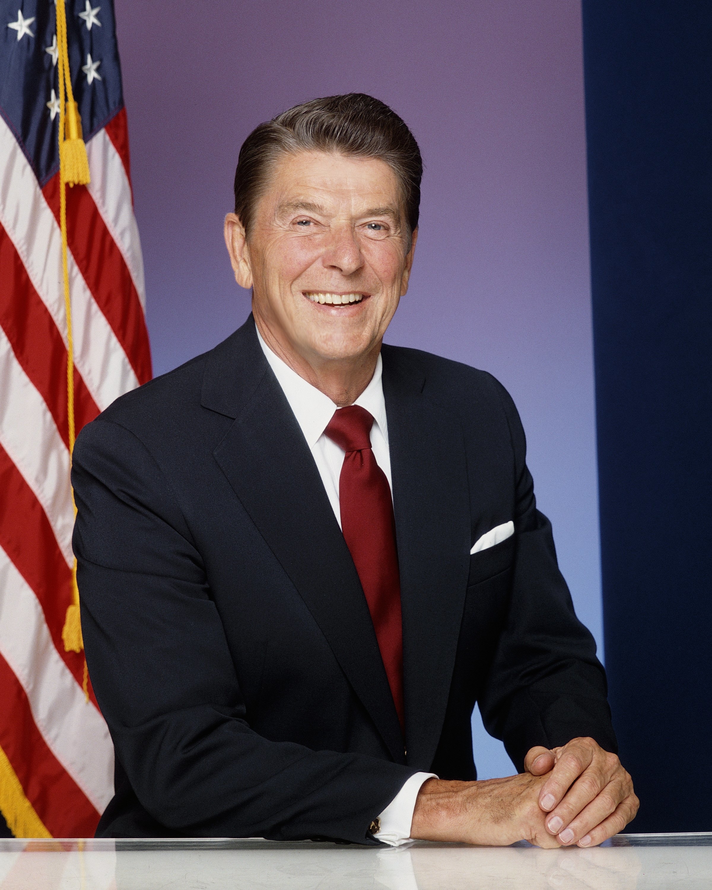Ronald Reagan, smiling, dressed in a suit with a red tie, an American flag in the background