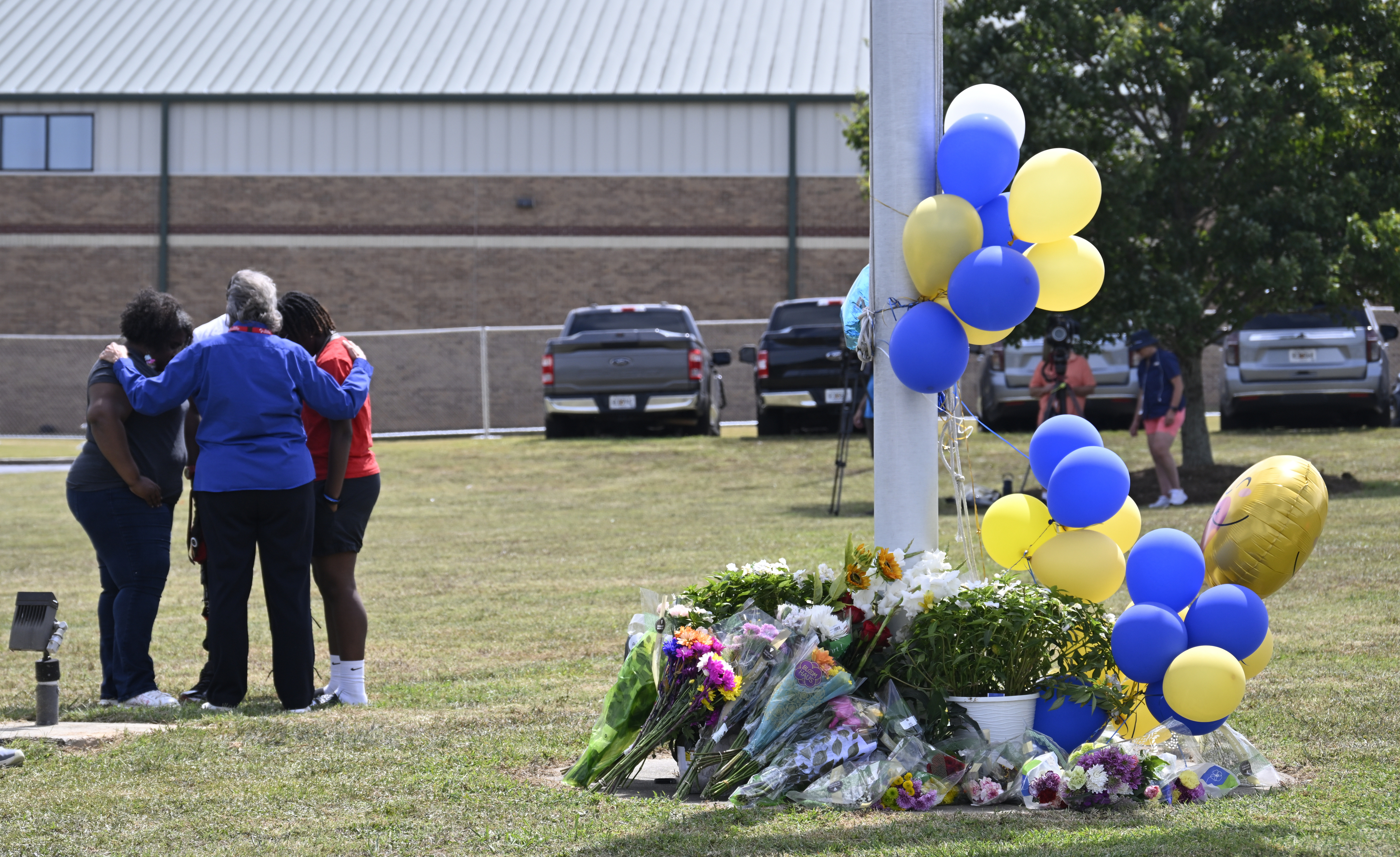 A small group of people embrace near a flagpole surrounded by flowers and blue and yellow balloons, likely commemorating a memorial or tribute event