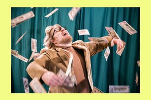 Person in a beige blazer and glasses, standing with arms raised, while money falls around them