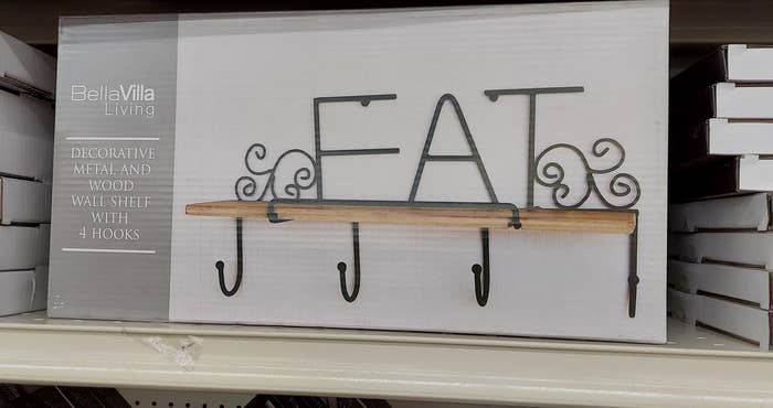 Decorative metal and wood wall shelf with 4 hooks, designed with the word "EAT" and swirl accents, displayed on a store shelf