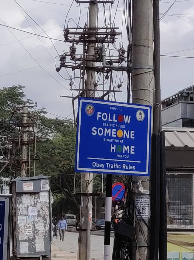 A blue road sign reads: "Follow traffic rules. Someone is waiting at home for you. Obey traffic rules."