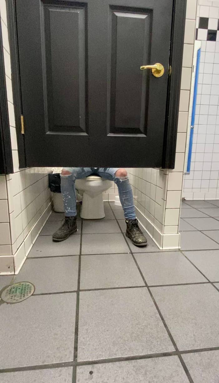A person in ripped jeans and boots is sitting in a bathroom stall, with their legs visible under the door