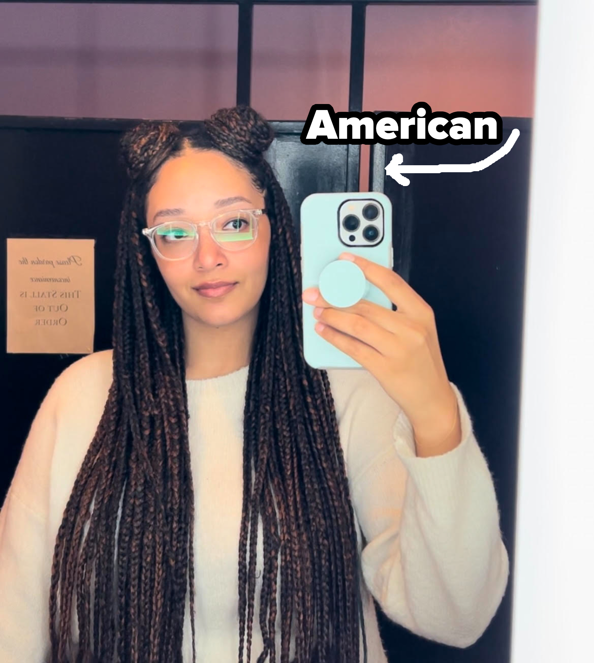 A woman with long braided hair and glasses takes a mirror selfie holding a smartphone with a pop socket. She wears a light sweater