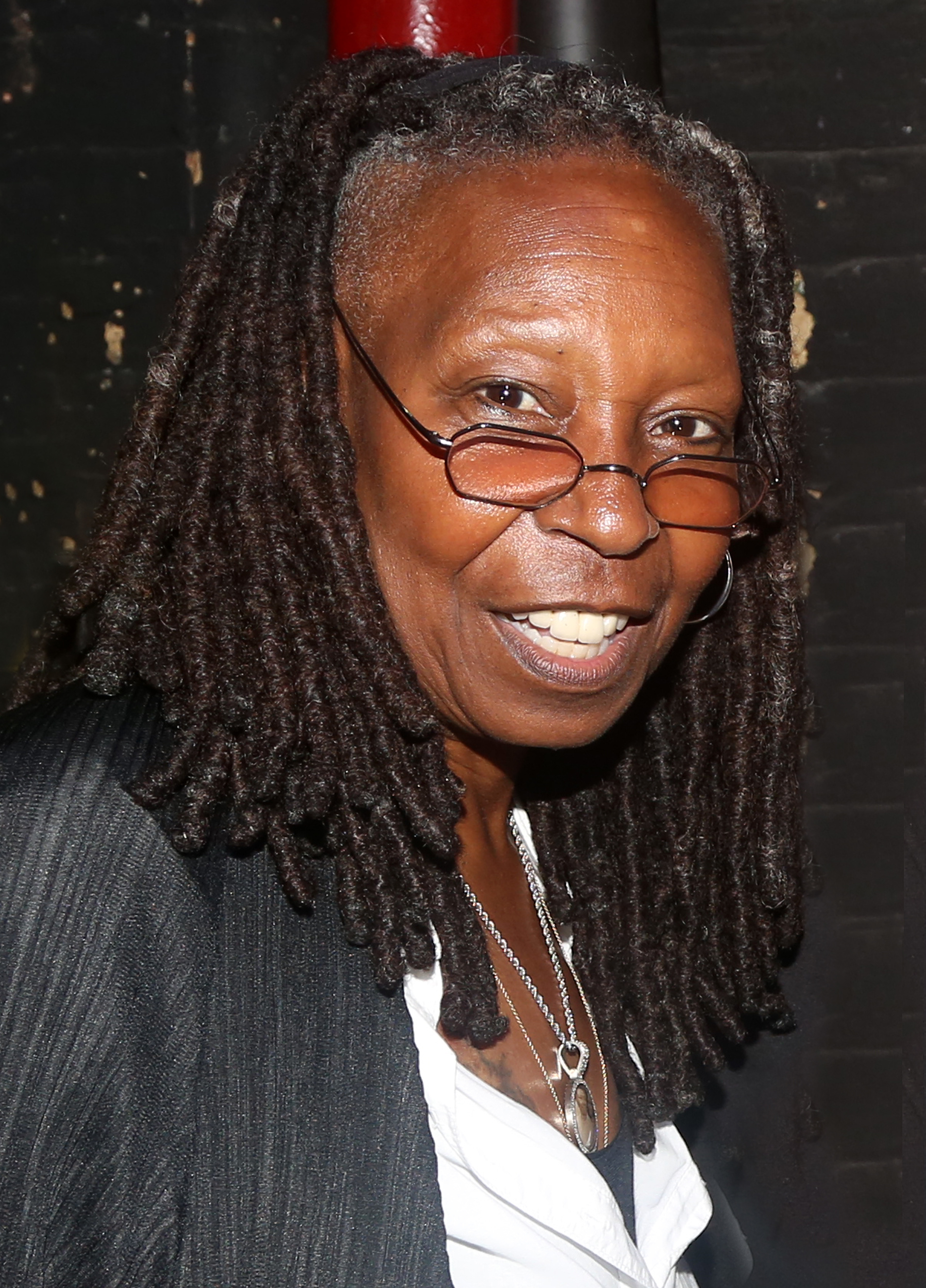 Whoopi Goldberg smiling, wearing glasses and a necklace over a white shirt with a black jacket
