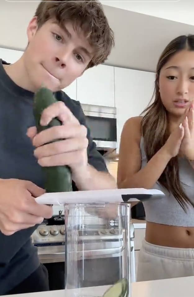 Two people prepare food in a kitchen. One is grating a zucchini over a container, while the other watches attentively