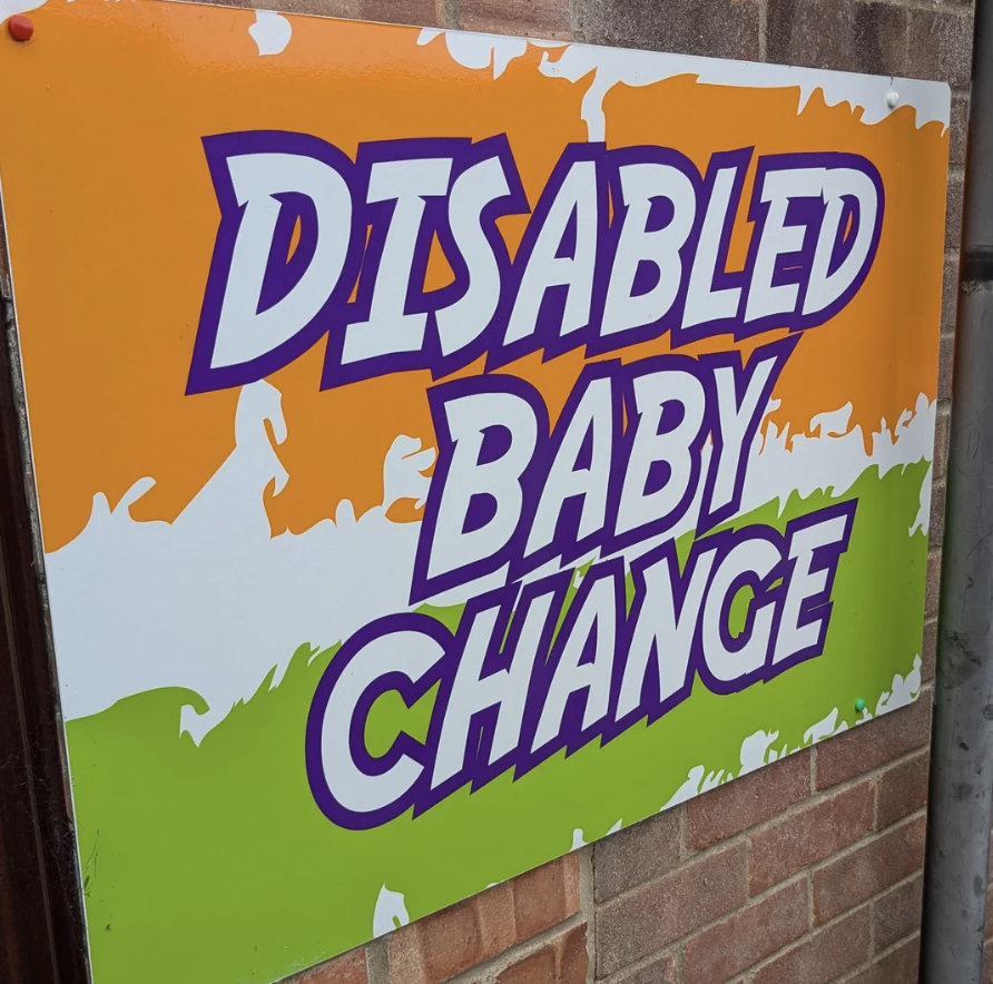 A sign reads "Disabled Baby Change" with text in bold, purple and white letters on an orange, white, and green background
