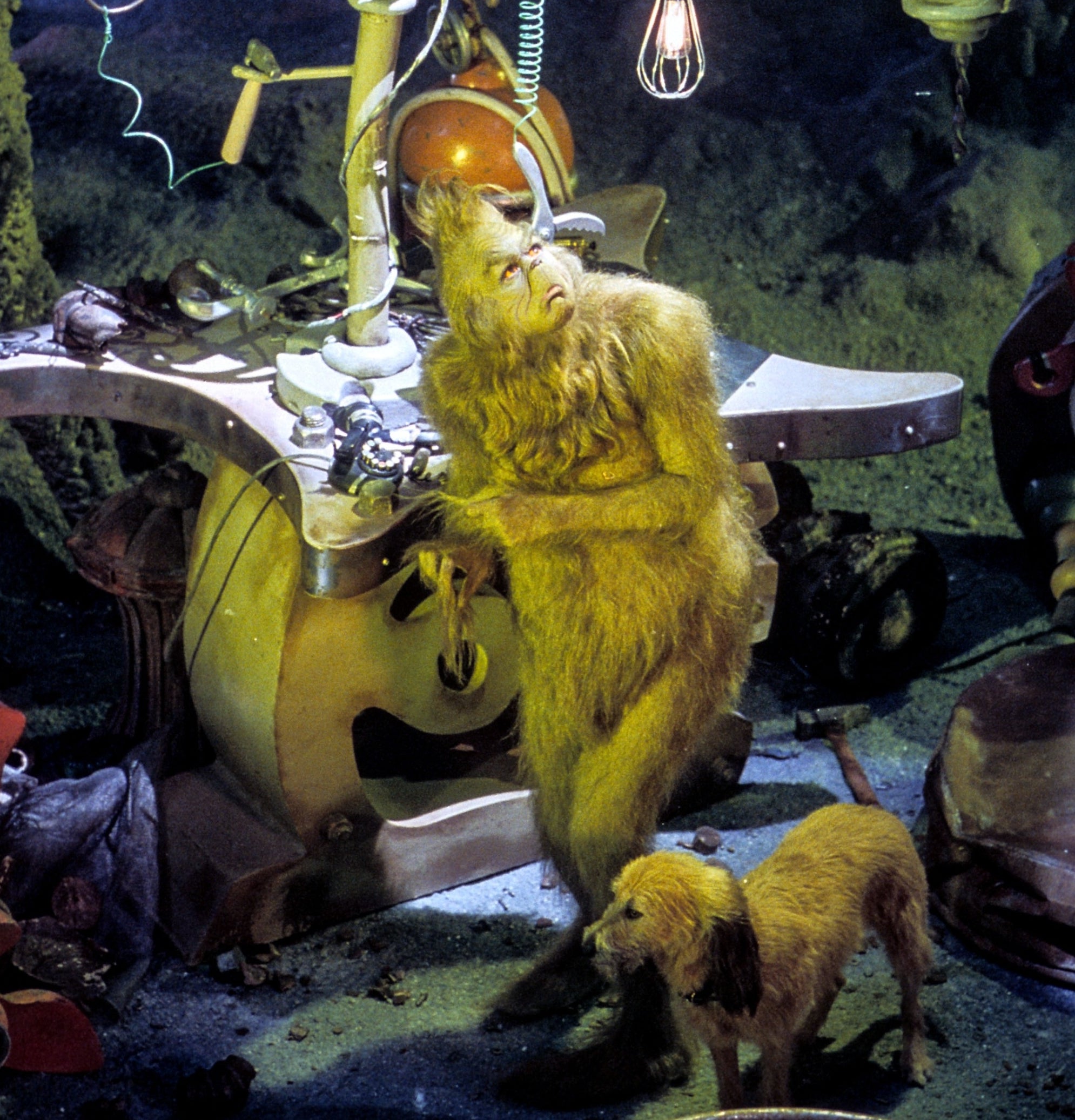 The Grinch from "How the Grinch Stole Christmas!" stands in his workshop, looking contemplative, with Max the dog beside him. Various inventive gadgets surround them
