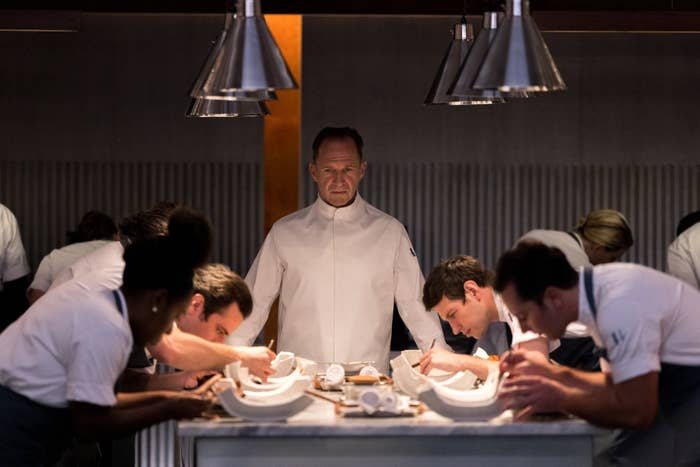 Ralph Fiennes in a chef's outfit overlooking a table with people working on fancy dishes in &quot;The Menu&quot;