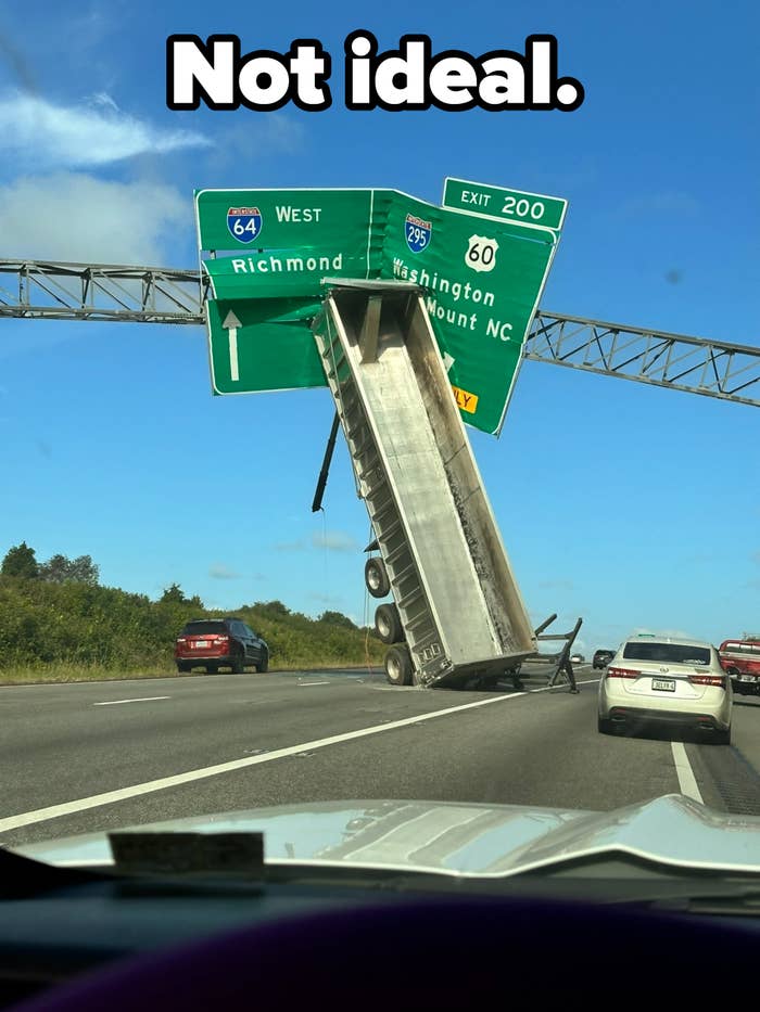A truck's trailer is lifted upright, stuck on a highway exit sign over an interstate, causing traffic to slow down around it