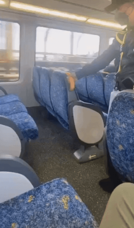 A conductor on a train uses a yellow tool to check under a blue passenger seat, ensuring safety onboard