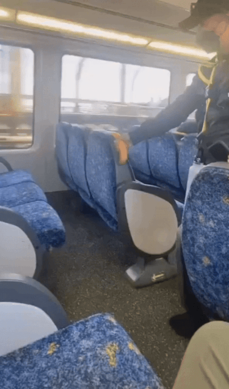 A conductor on a train uses a yellow tool to check under a blue passenger seat, ensuring safety onboard