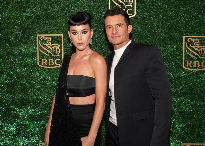 Katy Perry and Orlando Bloom pose together. Katy wears an elegant strapless dress and drop earrings. Orlando is in a sleek suit with an undershirt