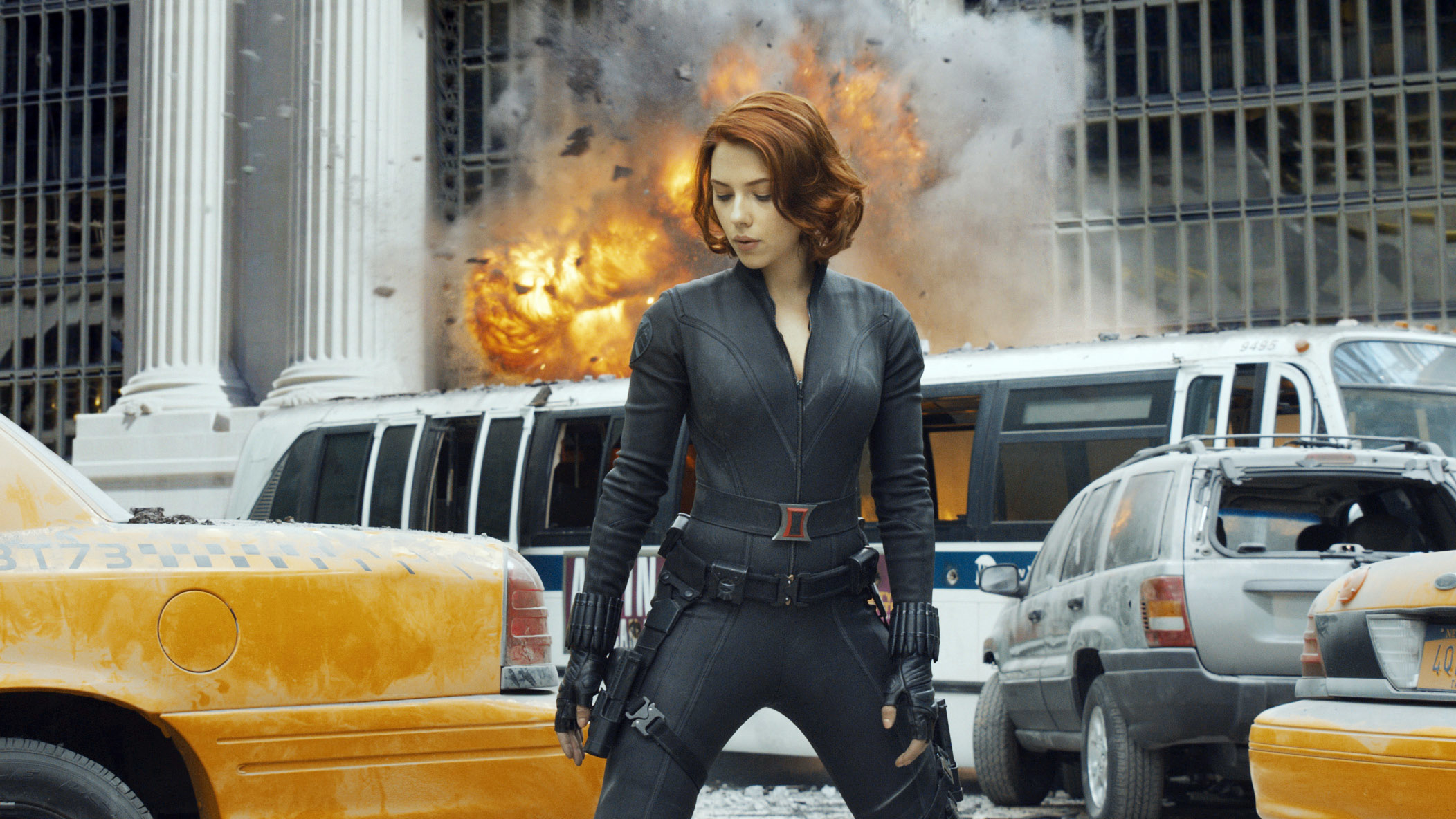 Scarlett Johansson as Black Widow in a tight tactical suit, stands amid debris and taxis with an explosion in the background