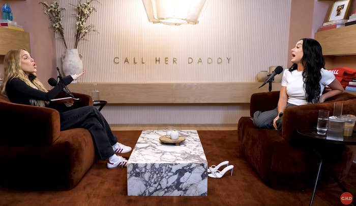 Alex Cooper and guest in an animated conversation on "Call Her Daddy" podcast. Both seated in armchairs with mics, table with decor in between
