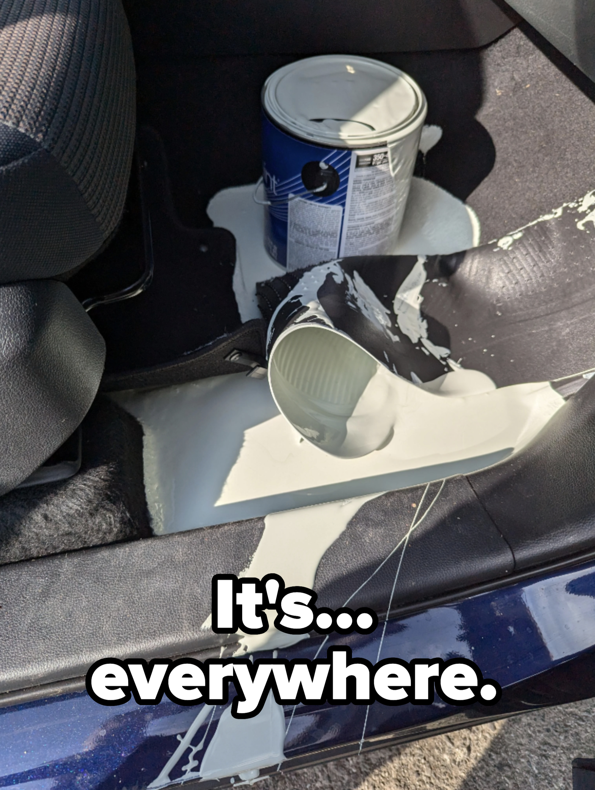 A spilled can of white paint on a car's floor, with the paint spreading across the floor mat and into the car seat