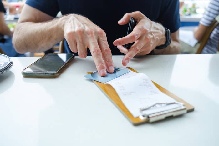 A person holds a credit card above a restaurant bill on a clipboard, with a smartphone on the table nearby