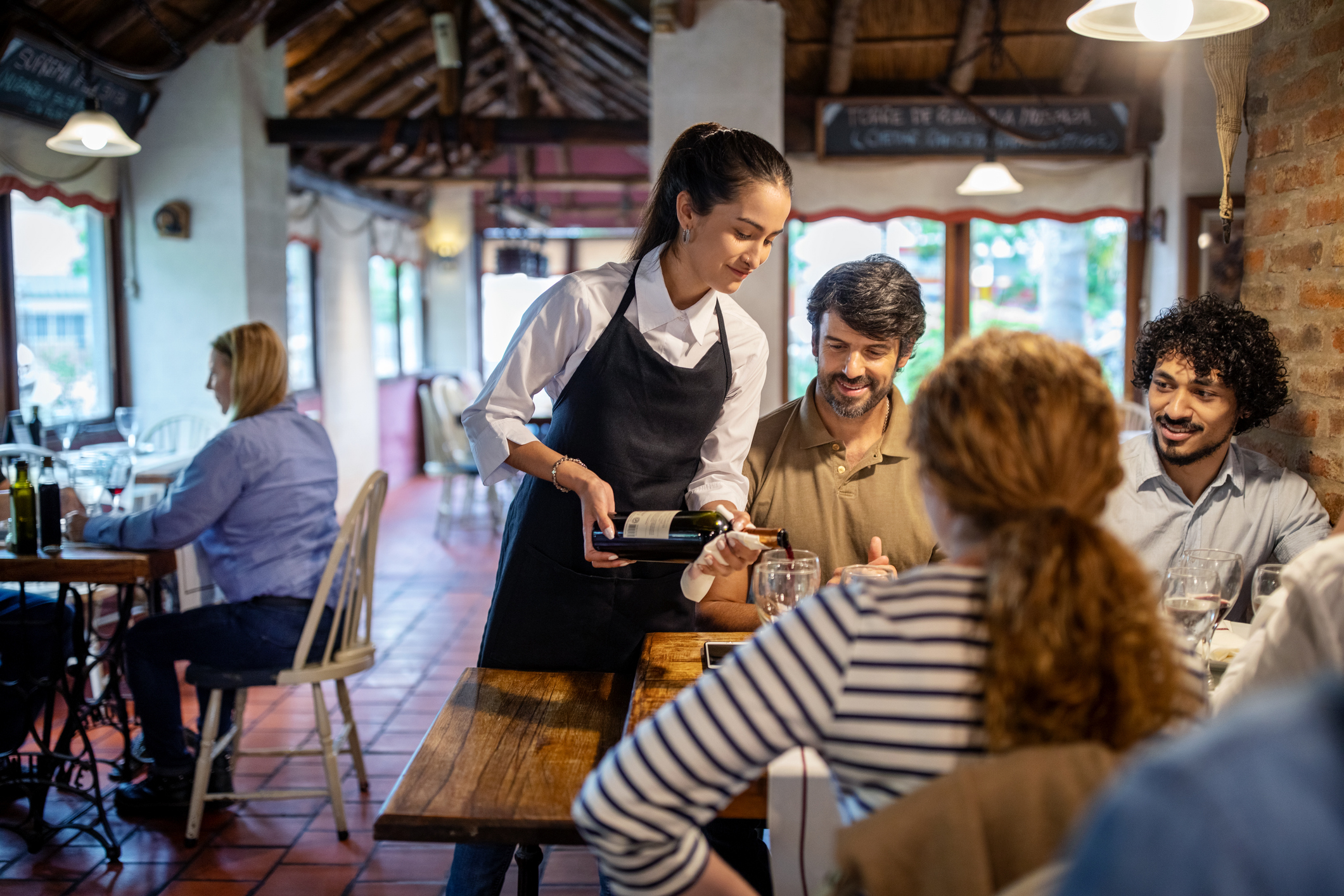 A waitress pours wine for two men, smiling at a restaurant table. Another woman and man sit at different tables in the background