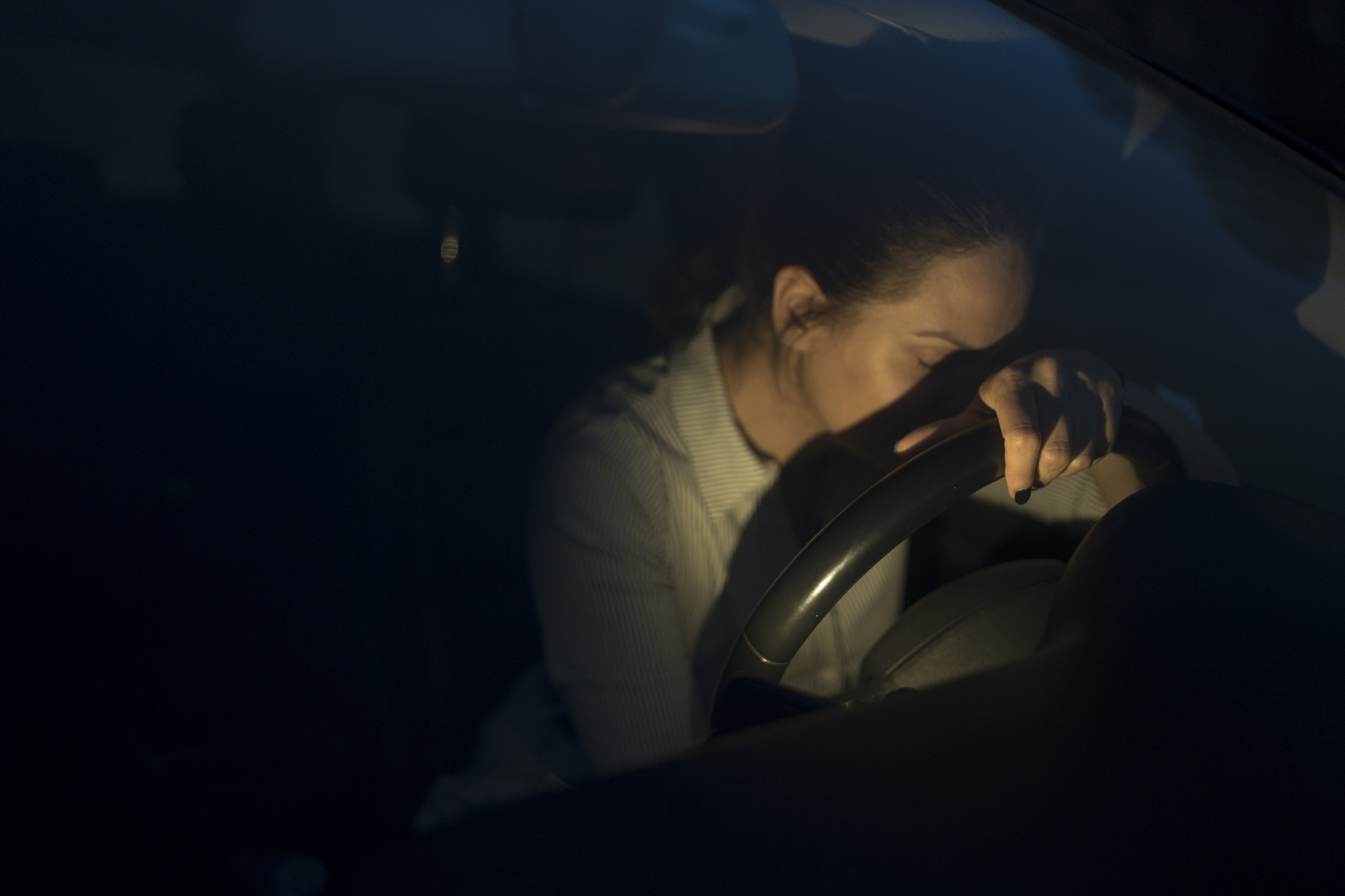 A person sits in a car at night, resting their head on the steering wheel, appearing tired or reflective