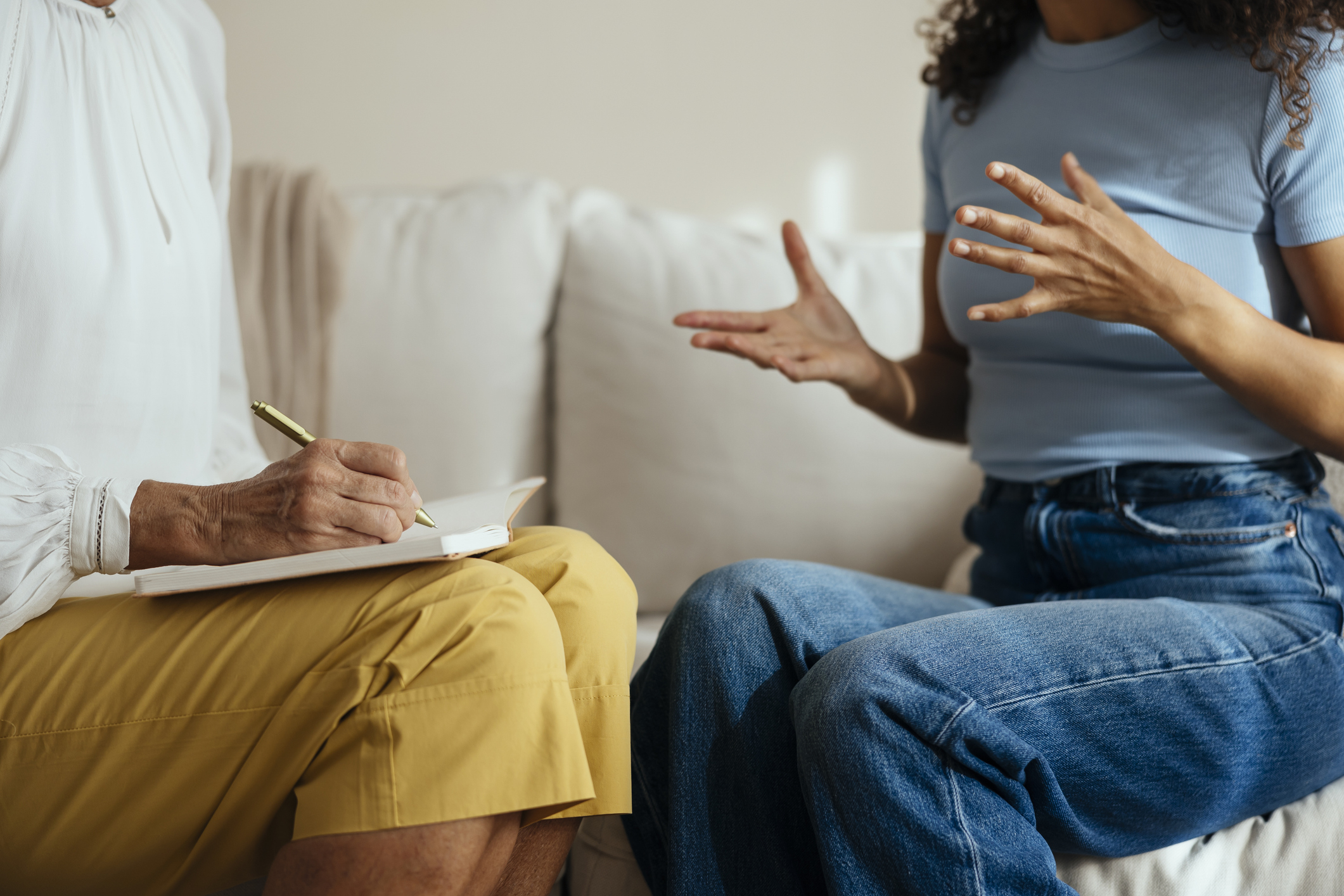 Two individuals seated on a couch are engaged in conversation, one is taking notes while the other is gesturing expressively