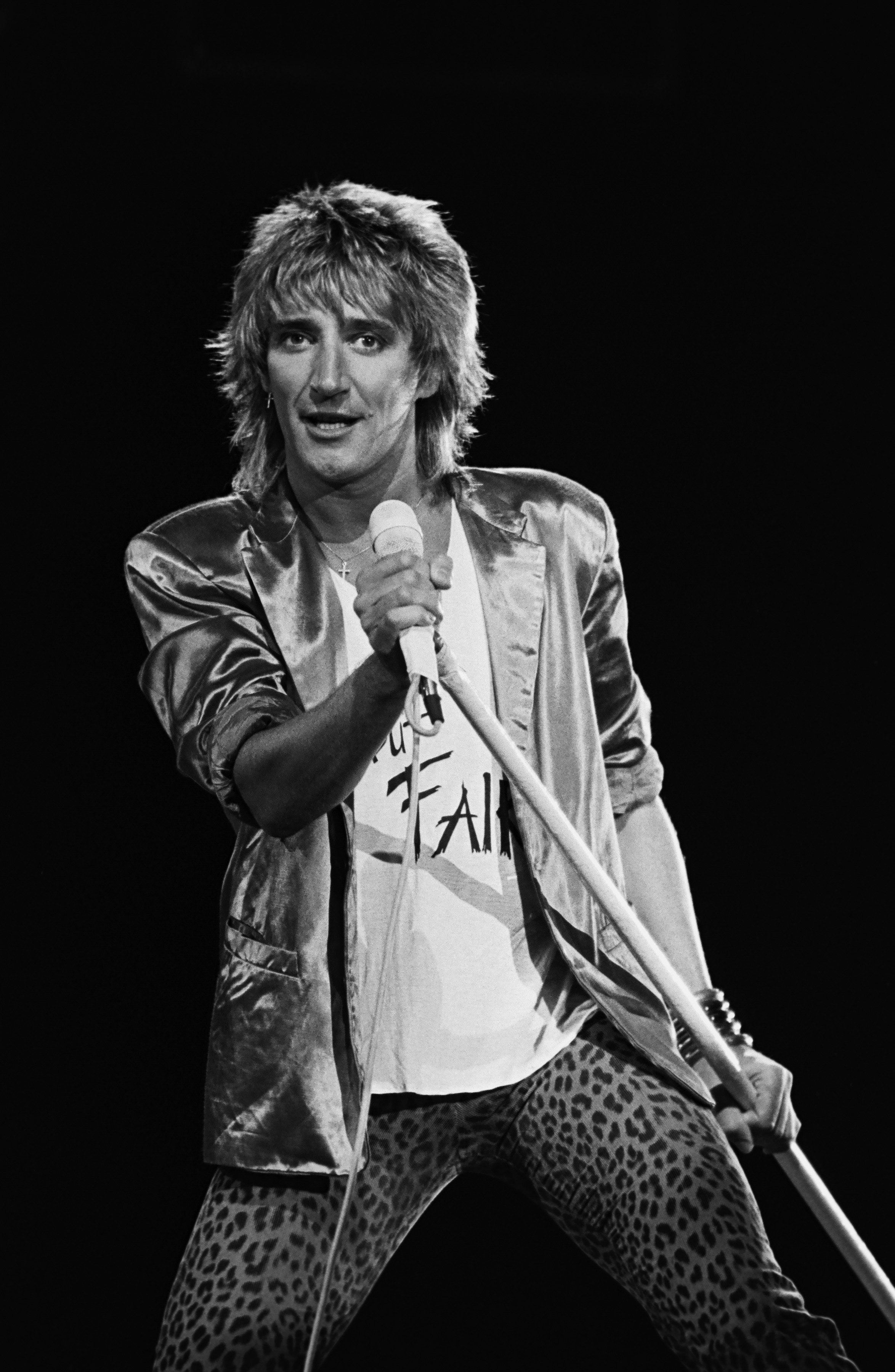 Rod Stewart performs on stage, wearing a shiny jacket and leopard print pants, while holding a microphone stand