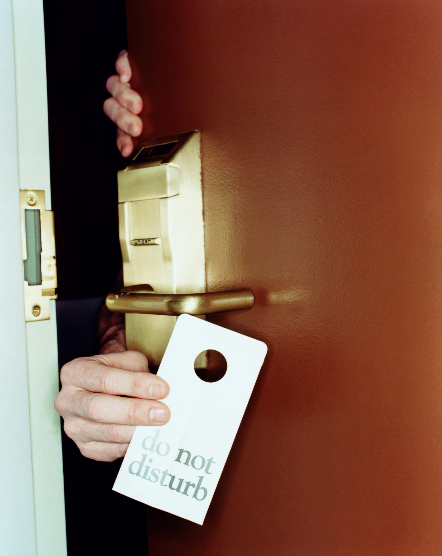 A hand is holding a "do not disturb" sign on a hotel door handle, with another hand seen behind the door