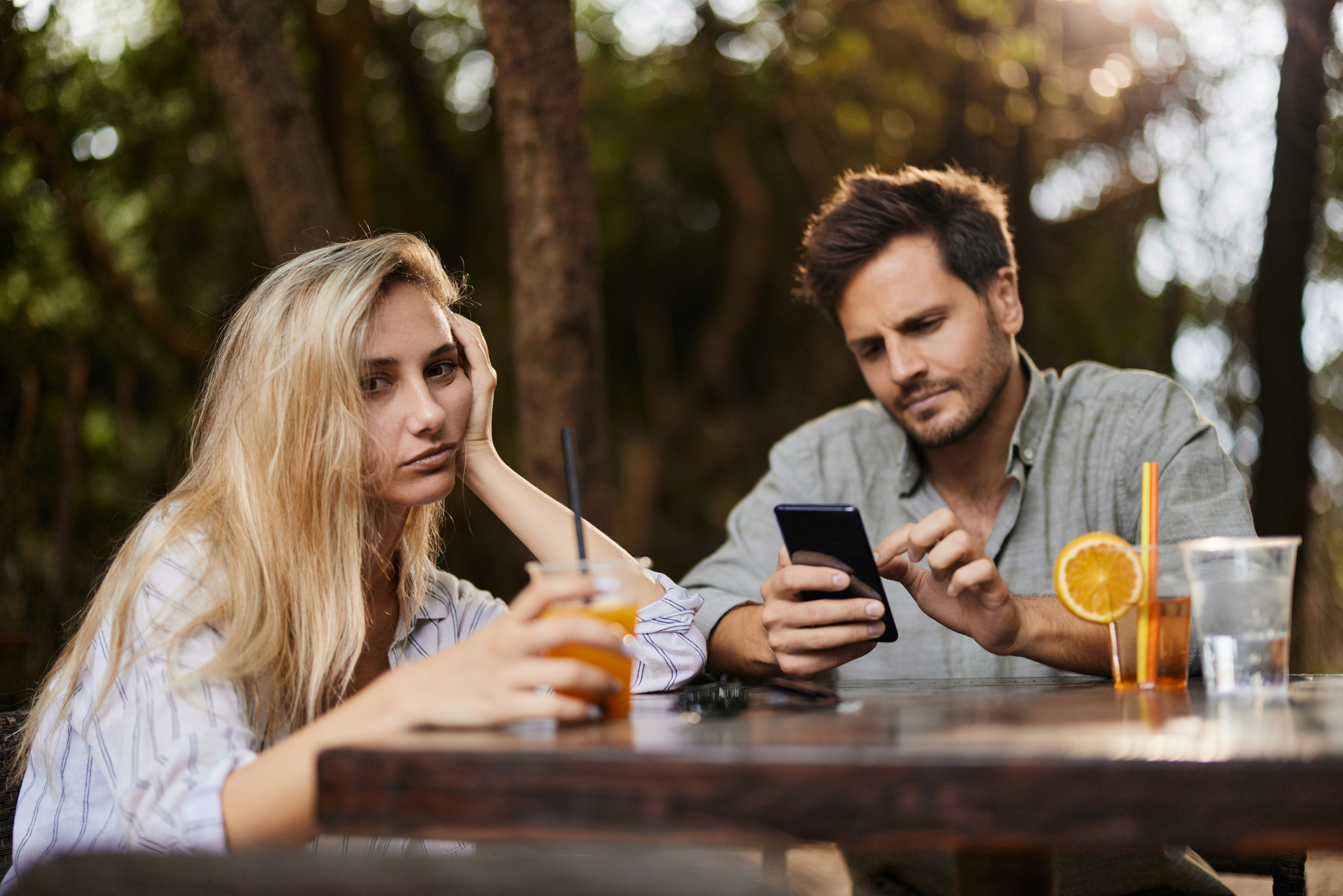 A woman looks bored while holding a drink, as a man beside her focuses on his phone at an outdoor table