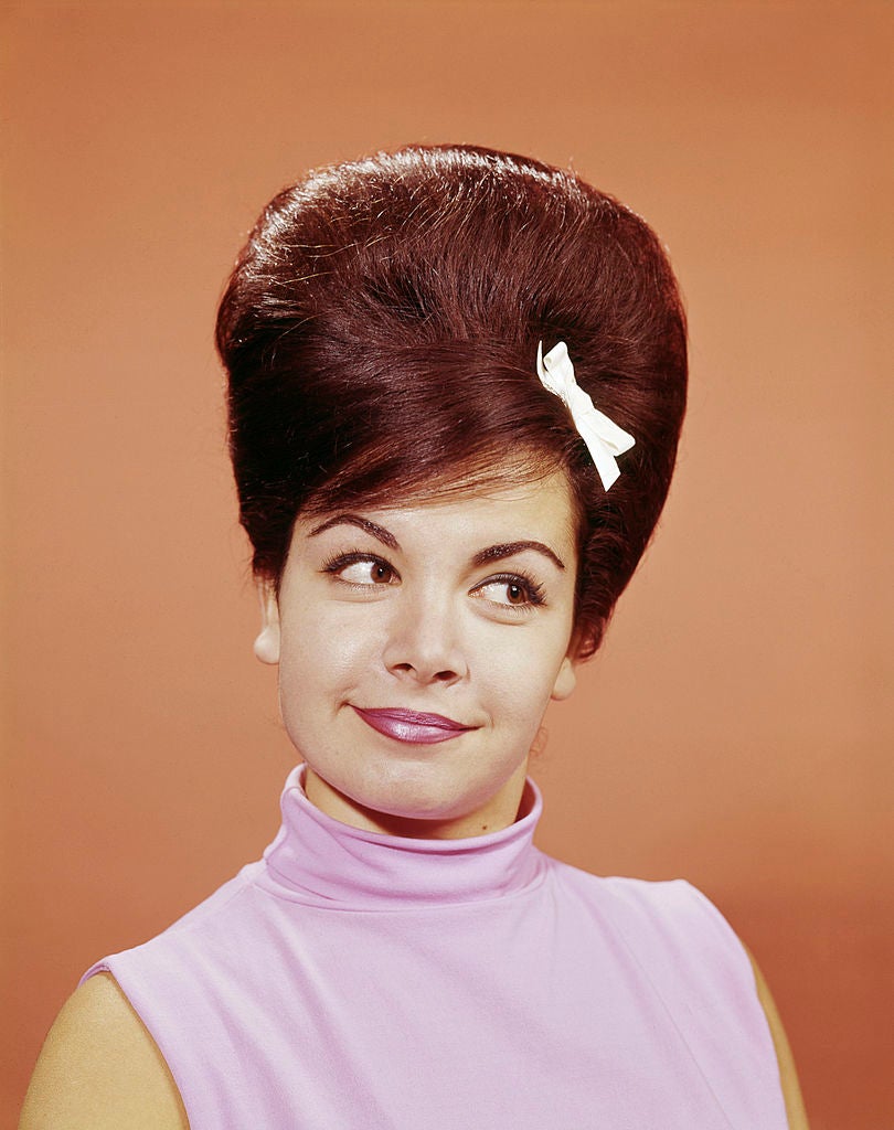 A woman with a beehive hairstyle and a white bow, wearing a sleeveless high-neck top, looks off to the side with a subtle smile