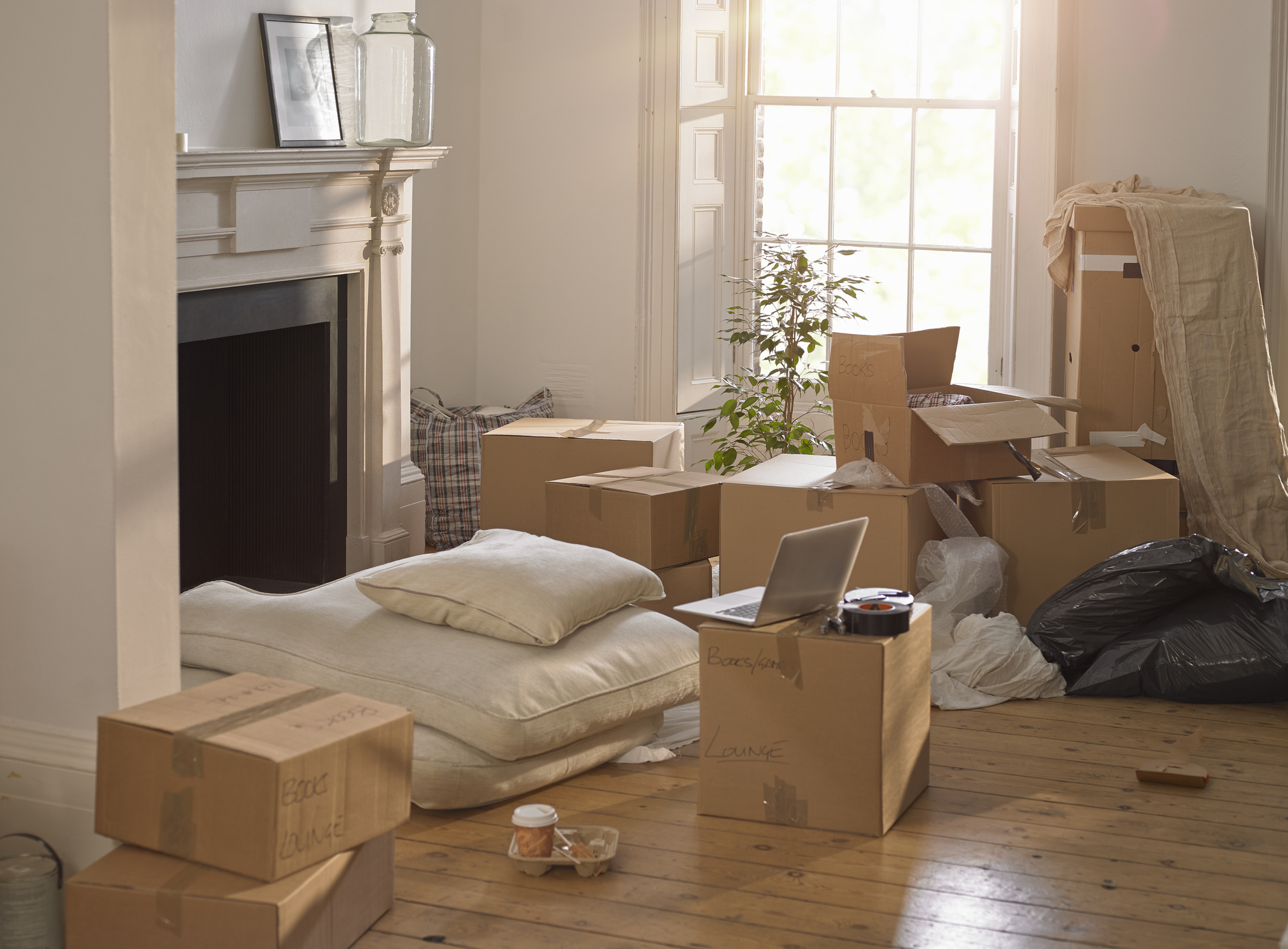 A sunlit room with packed moving boxes, a disassembled couch, and a laptop on a box, suggesting a couple's new beginning or moving in together
