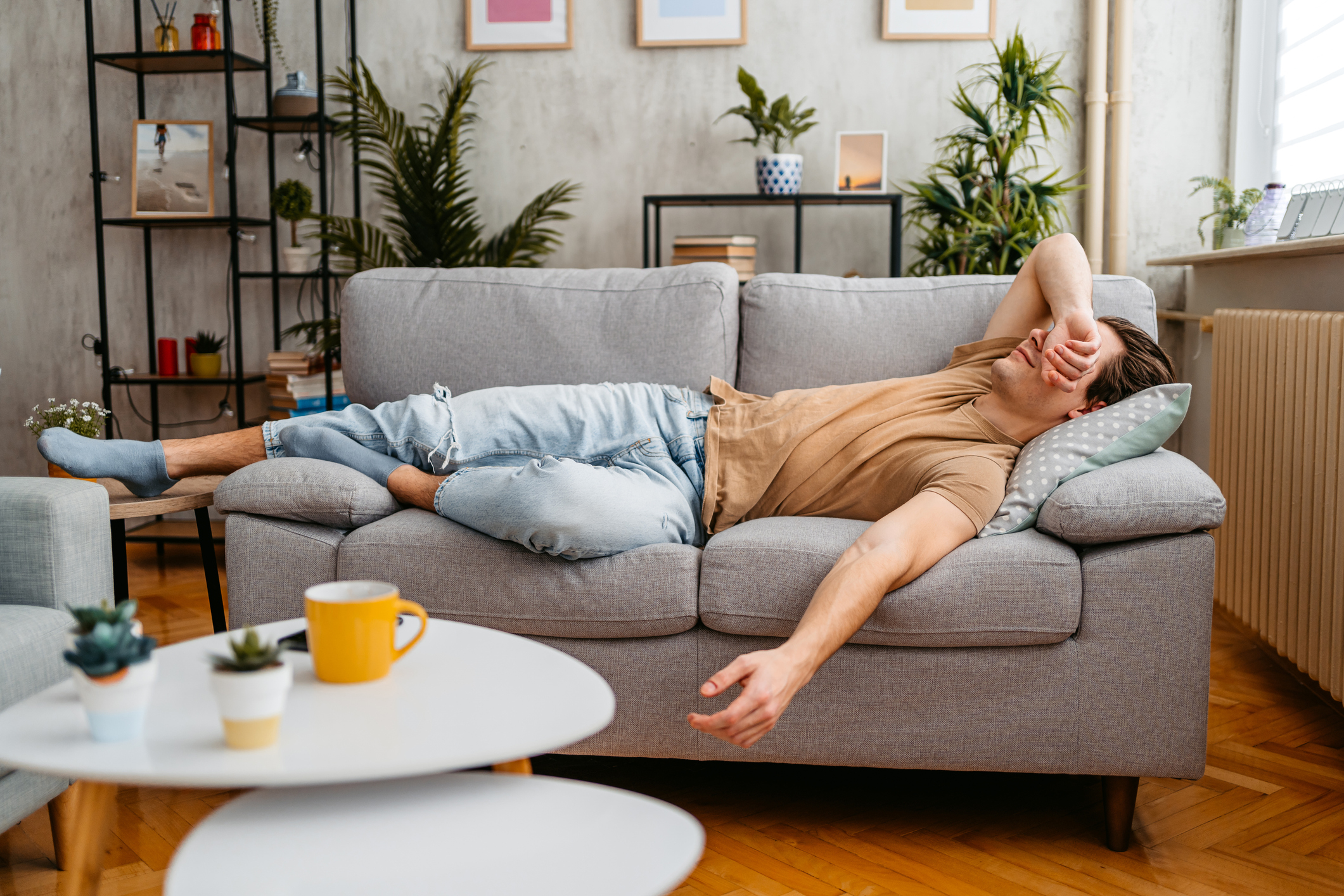 A person wearing a casual short-sleeve shirt and ripped jeans is lying on a couch, covering their face with their arm in apparent fatigue