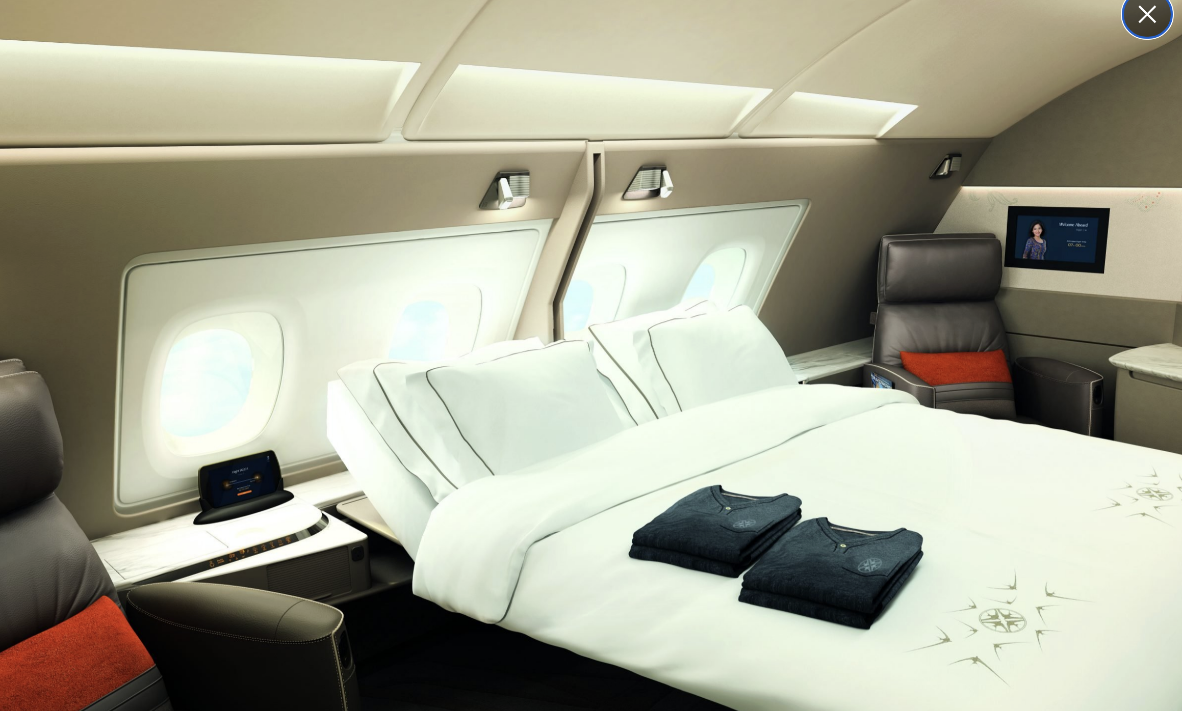 Luxury first-class airplane suite with a double bed, leather armchairs, fold-out TV, and amenity kits on the bed