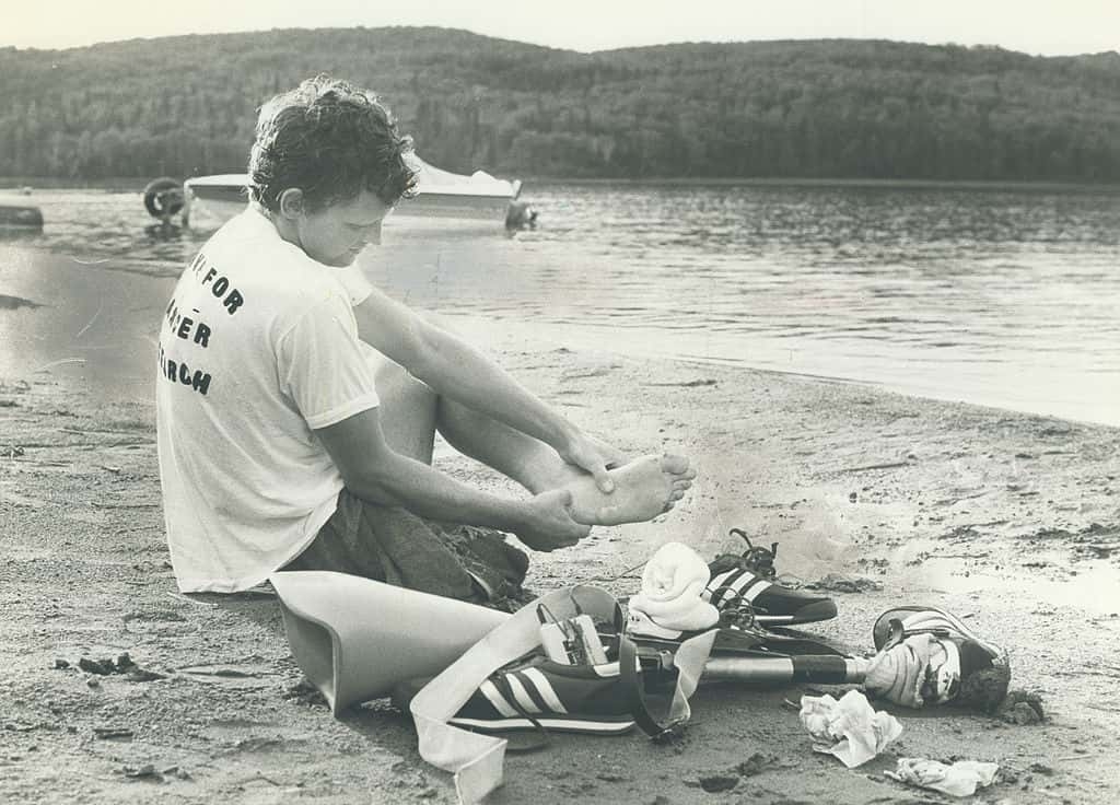 Terry Fox sits on a lakeshore, massaging his left foot, with a prosthetic leg, a pair of Adidas shoes, and some items placed on the sand beside him