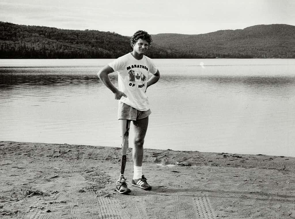 Terry Fox stands on a sandy shore with a prosthetic leg, wearing athletic shorts, a "Marathon of Hope" T-shirt, and sneakers, with a lake and forested hills behind him