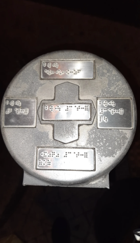 Metallic circular surface with four Braille inscriptions facing different directions. The inscriptions are likely tactile guidance points on an urban fixture