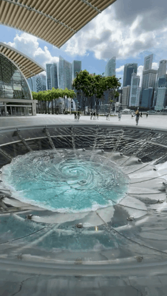 A large urban water vortex installation next to a modern building with a backdrop of skyscrapers and trees