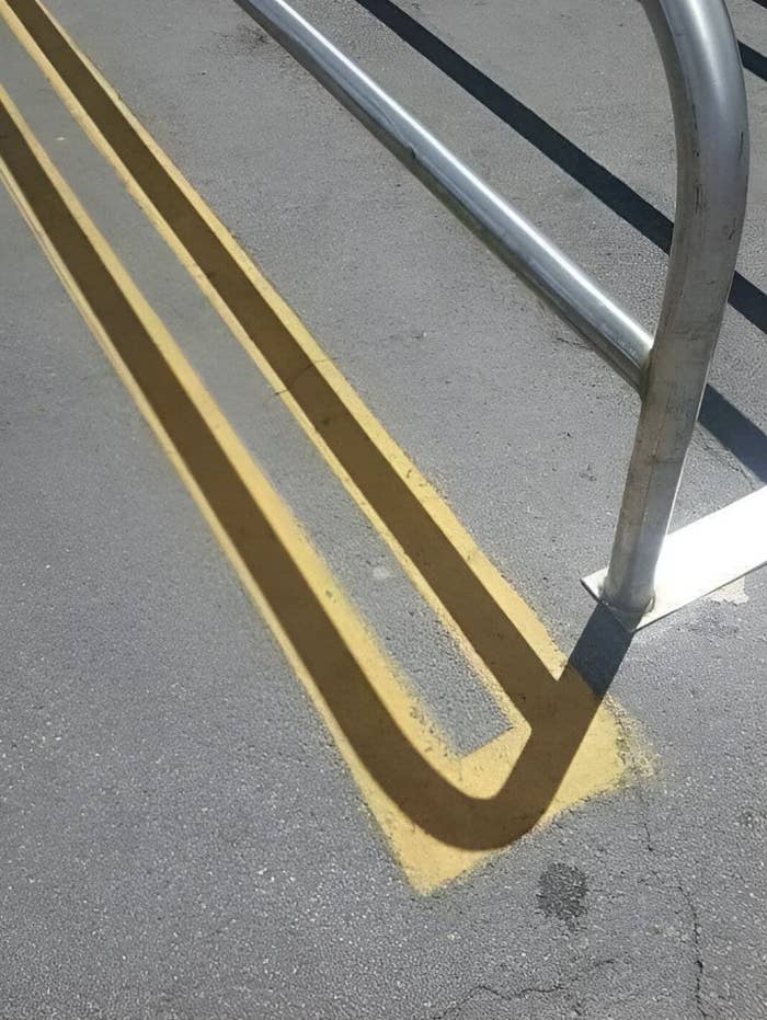 A shadow of a bike rack forms an optical illusion resembling a 2-dimensional drawing of a 3-dimensional object on a paved surface