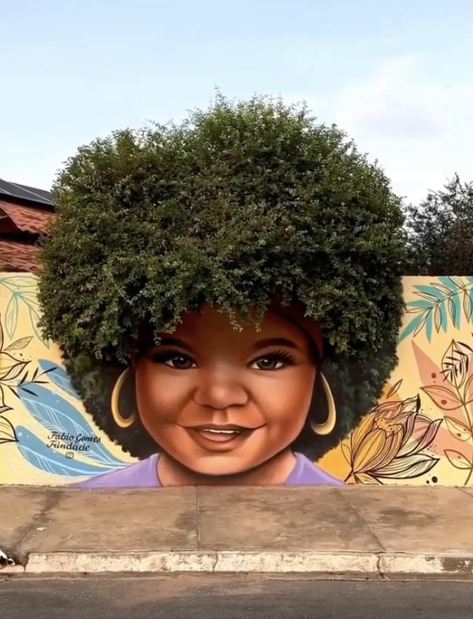 Mural of a smiling child with gold hoop earrings, brown skin, and an afro cleverly depicted using a tree. Text on mural: "Fábio Gomes Trindade"