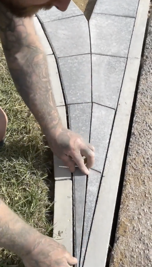 A person with tattooed arms is showing a method of arranging triangular and rectangular concrete pavers on a pathway near a grassy area