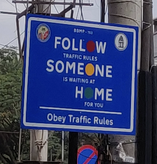 A blue road sign reads "FOLLOW TRAFFIC RULES - SOMEONE IS WAITING AT HOME FOR YOU. Obey Traffic Rules"