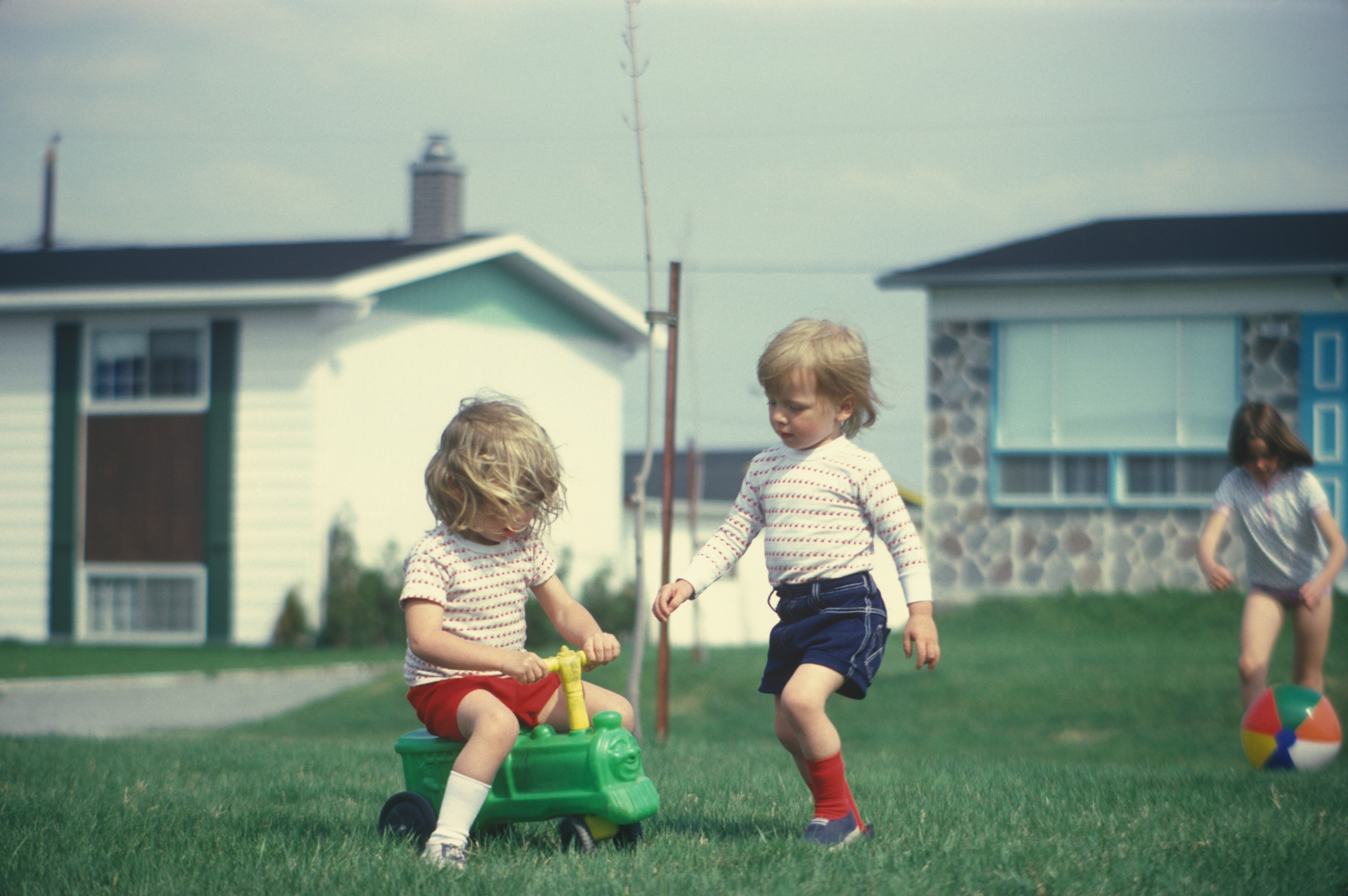 Two children play in a yard beside a small house, one on a toy vehicle and the other standing next to them. Another child plays with a ball in the background